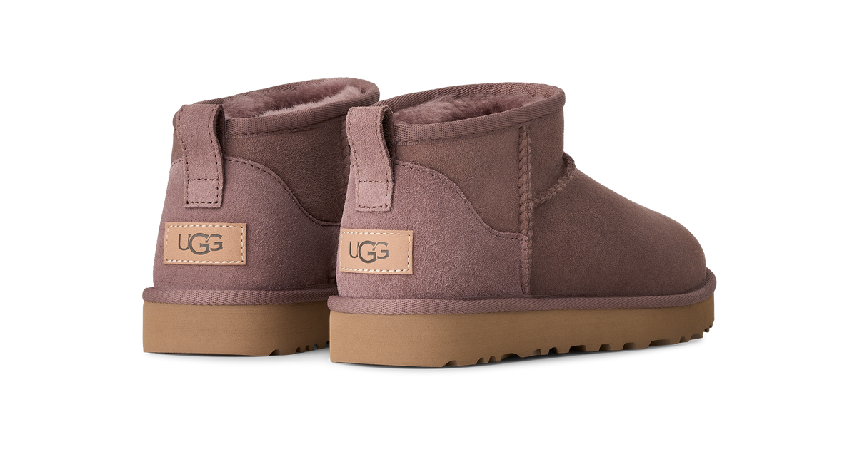 A pair of UGG mini boots in a dusty mauve-brown suede, featuring a tan leather UGG logo patch on the heel and a brown rubber sole, shown against a plain white background.