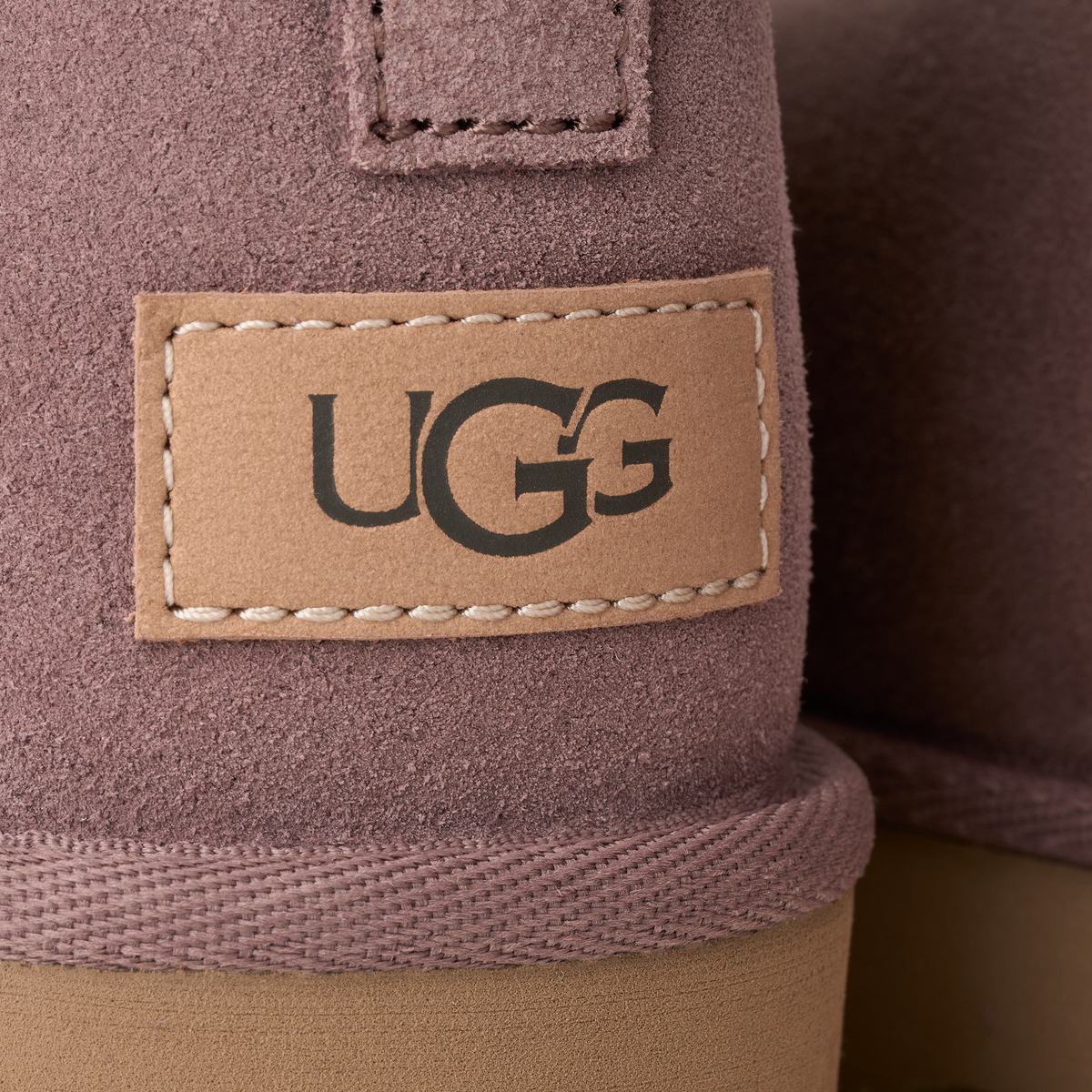 Close-up of a UGG boot showing the brand patch on a dusty mauve suede upper with a tan rubber sole.