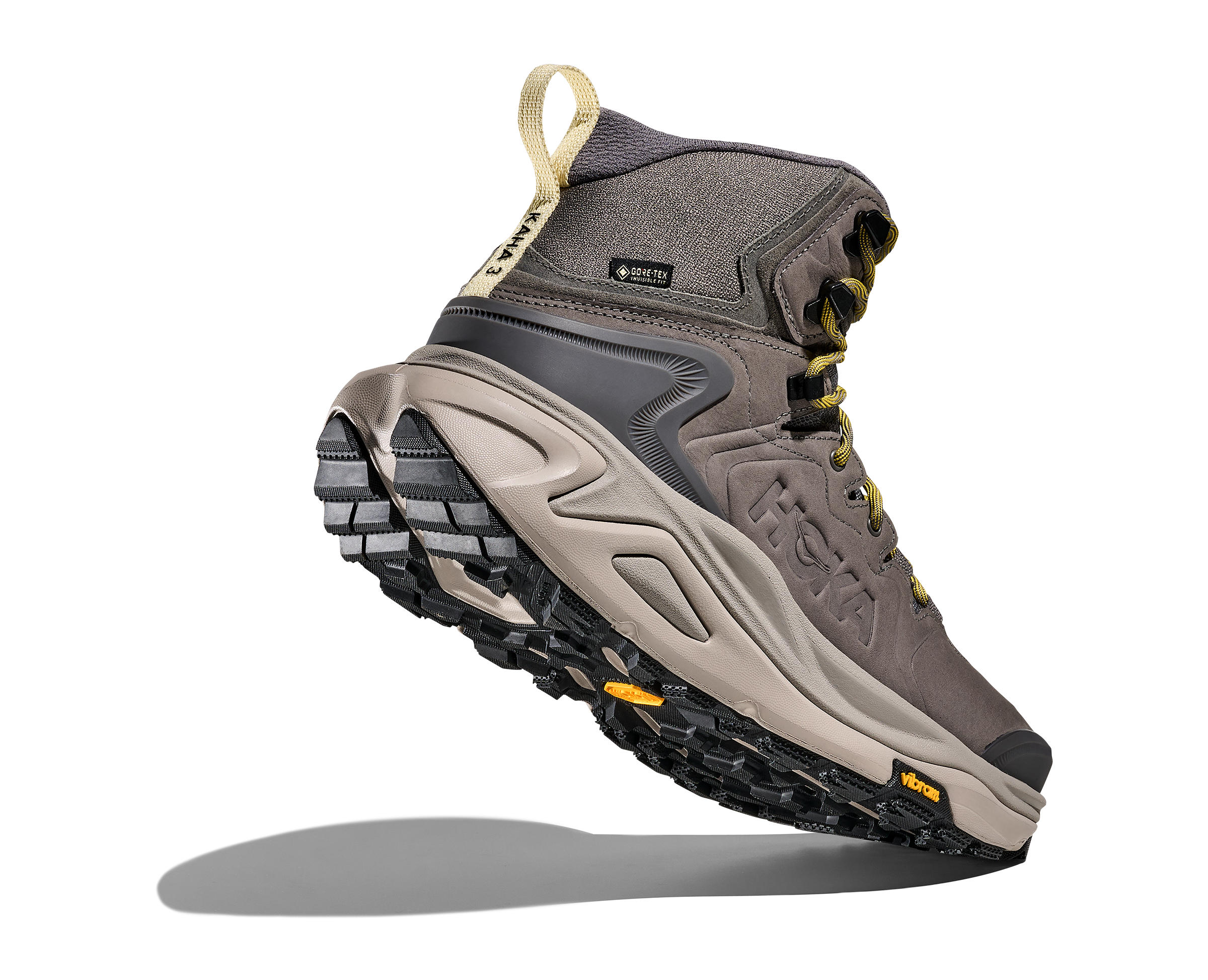 A single HOKA mid-cut hiking boot floating at an angle against a solid white background, showing its gray and beige upper, yellow laces, and black Vibram sole.
