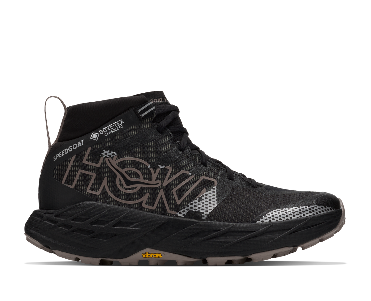 A black Hoka mid-cut trail running shoe with a textured mesh upper, featuring a large metallic bronze 'HOKA' logo on the side, a thick black and gray cushioned midsole, and a small yellow Vibram logo on the outsole.