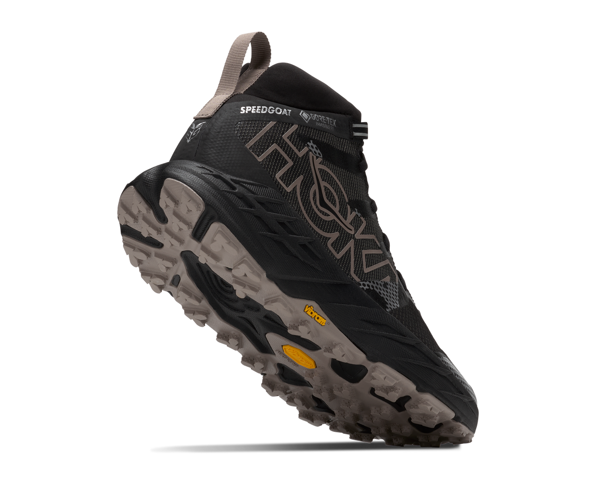 A Hoka Speed Goat mid-cut trail running shoe in black and dark gray with taupe accents and a yellow Vibram sole insert, shown on a clean white background.