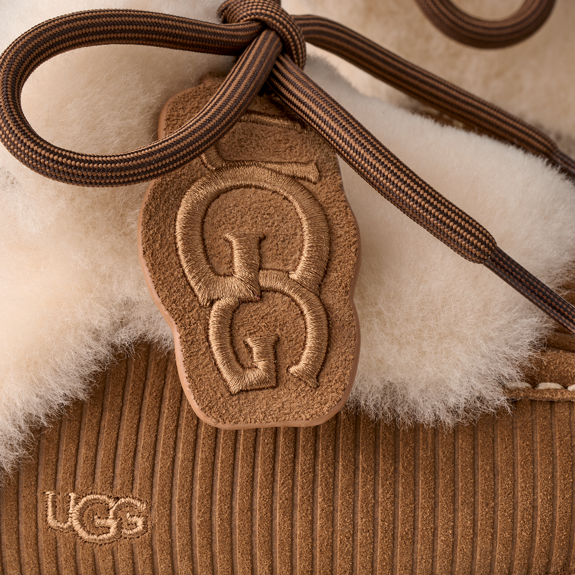 Close-up of a brown suede UGG pouch with a white shearling lining, featuring a matching brown leather UGG logo charm attached to a braided brown strap.