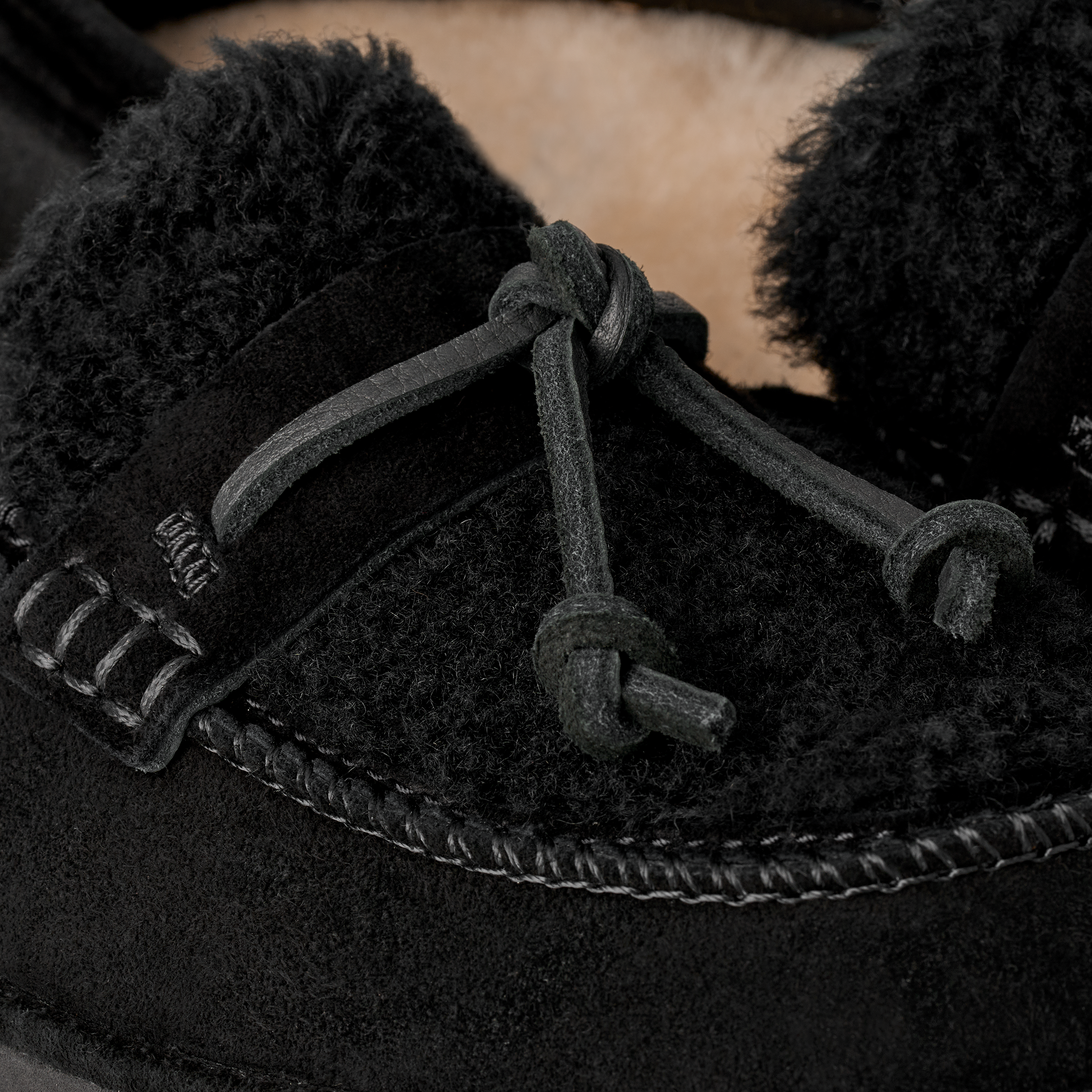 Close-up of a black suede moccasin-style boot with thick black shearling lining and knotted laces, shown against a soft beige background.
