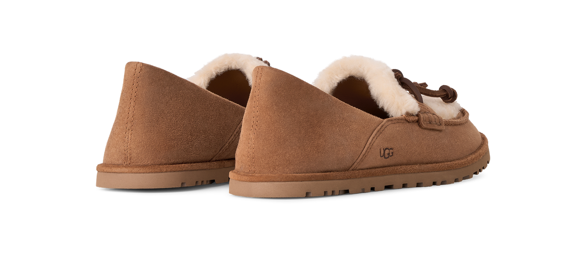 A pair of UGG slip-on suede slippers in a warm tan brown, featuring a soft cream shearling lining and a brown braided trim detail on the side, shown against a plain white background.