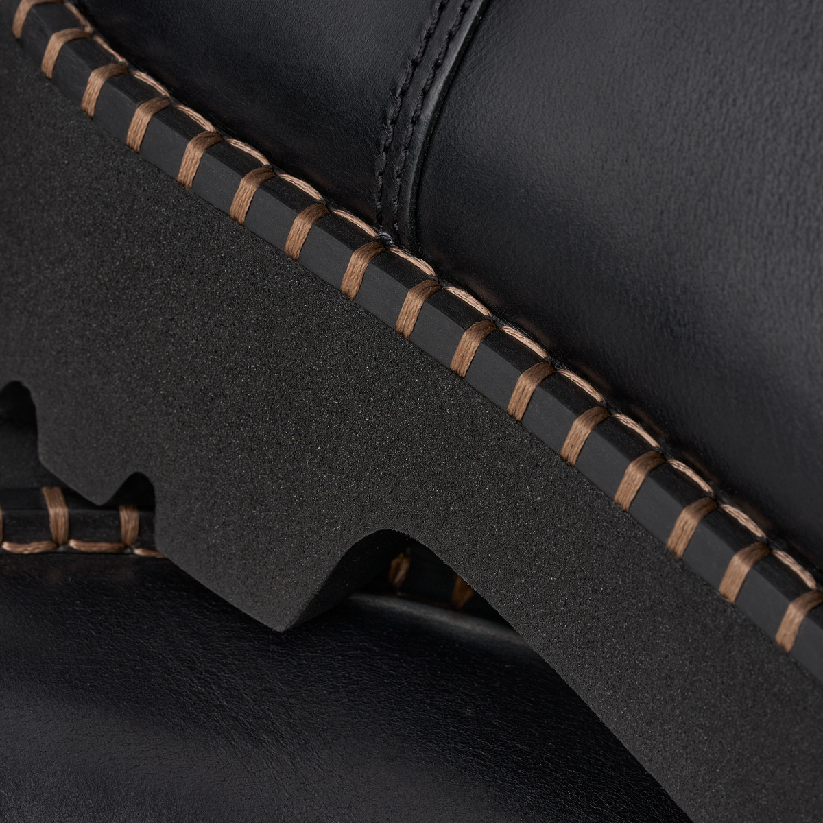 Close-up detail of a black leather boot showing the thick textured sole, contrast stitching, and layered edge construction.