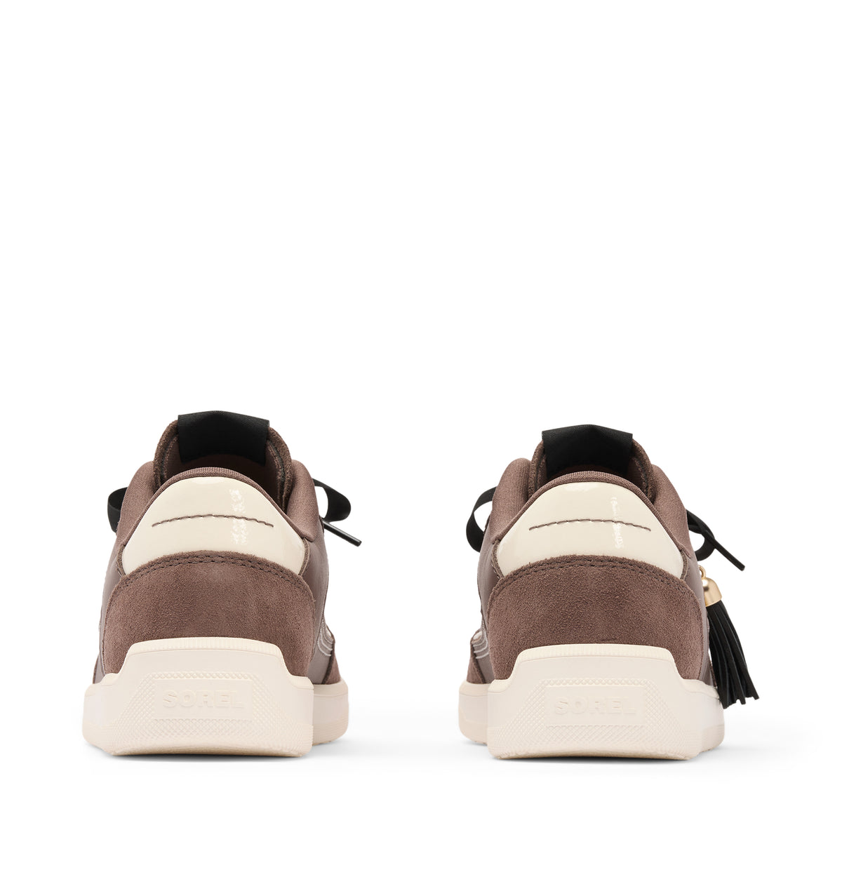A pair of brown suede and leather sneakers shown from the back, featuring cream-colored midsoles and heel tabs, black laces, and a black tassel with a gold charm on the right shoe.