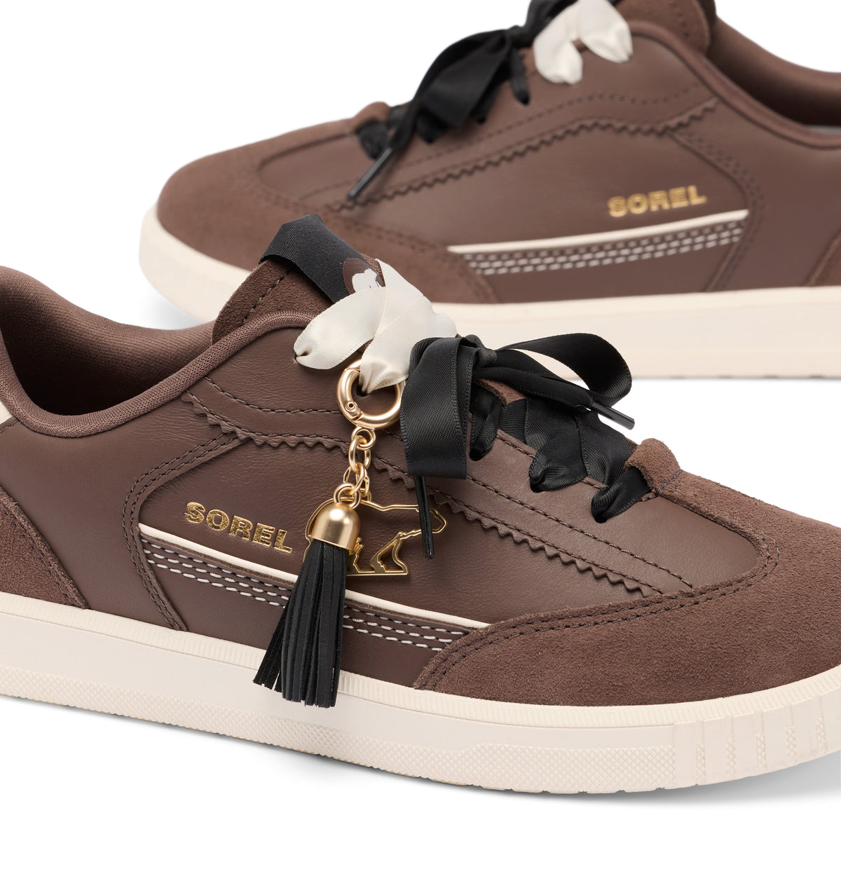 A pair of brown suede and leather low-top sneakers by Sorel, featuring black ribbon laces, a white horse-shaped charm, a black tassel, and a gold horse logo charm on the side.