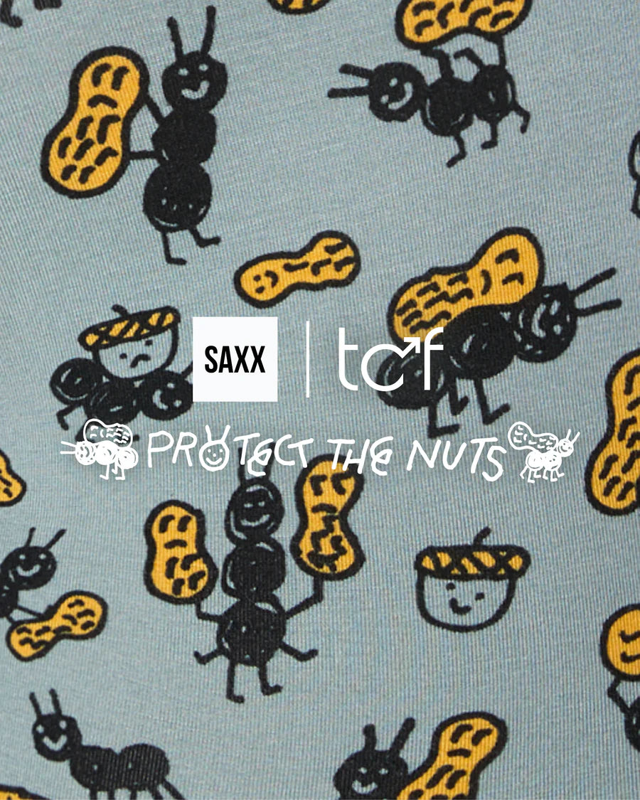 A close-up of a fabric pattern featuring cartoon ants carrying yellow peanuts on their backs, with a central white logo overlay reading 'SAXX | tcf PROTECT THE NUTS'.