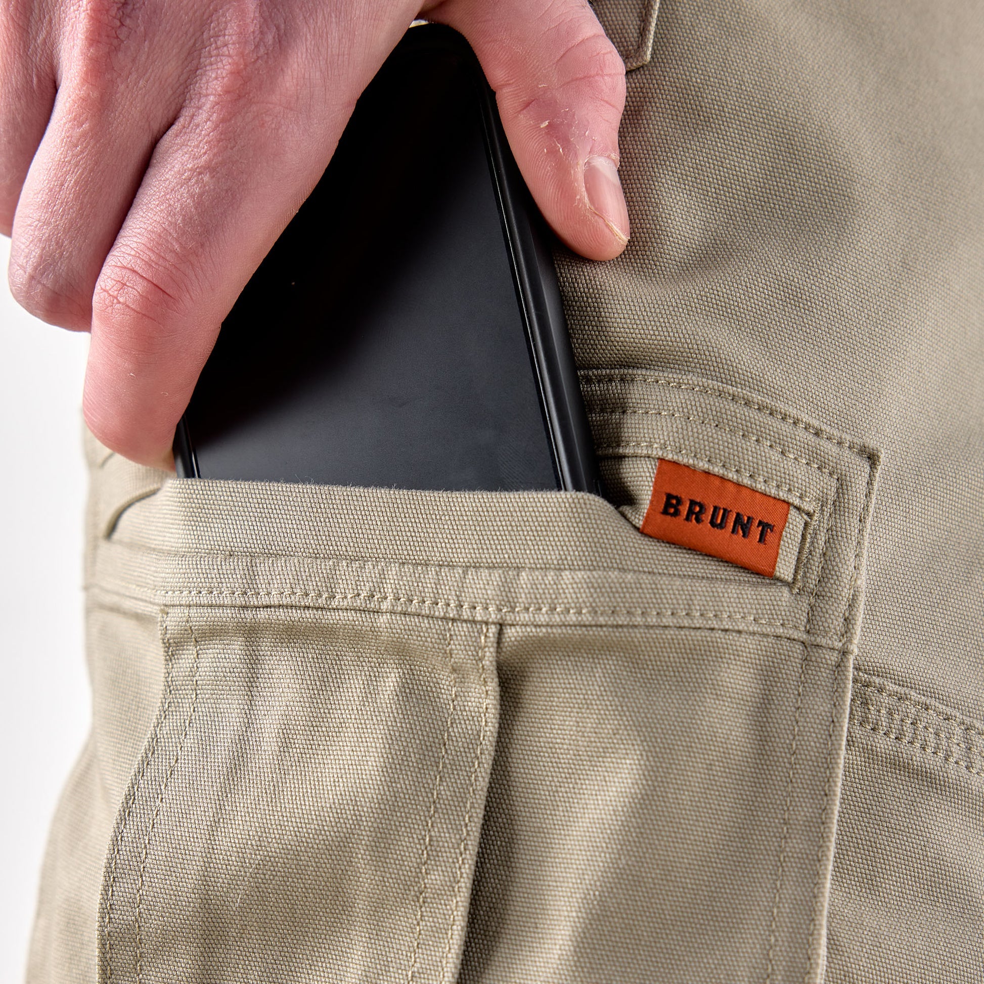 Person placing a phone into a pocket of beige pants with 'BRUNT' label.