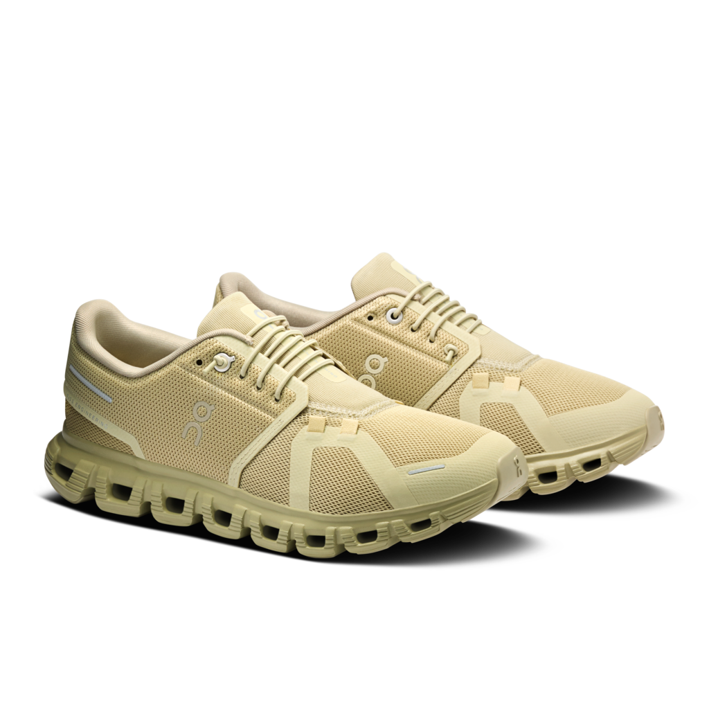 A pair of On running shoes in a light beige and cream colorway, featuring a chunky, segmented sole and the brand's logo on the side, set against a plain white background.