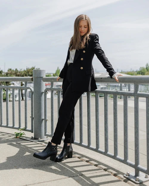 A woman stands outdoors leaning on a metal railing, wearing a black tailored blazer and matching trousers with gold-tone buttons, a white top, and black ankle boots.