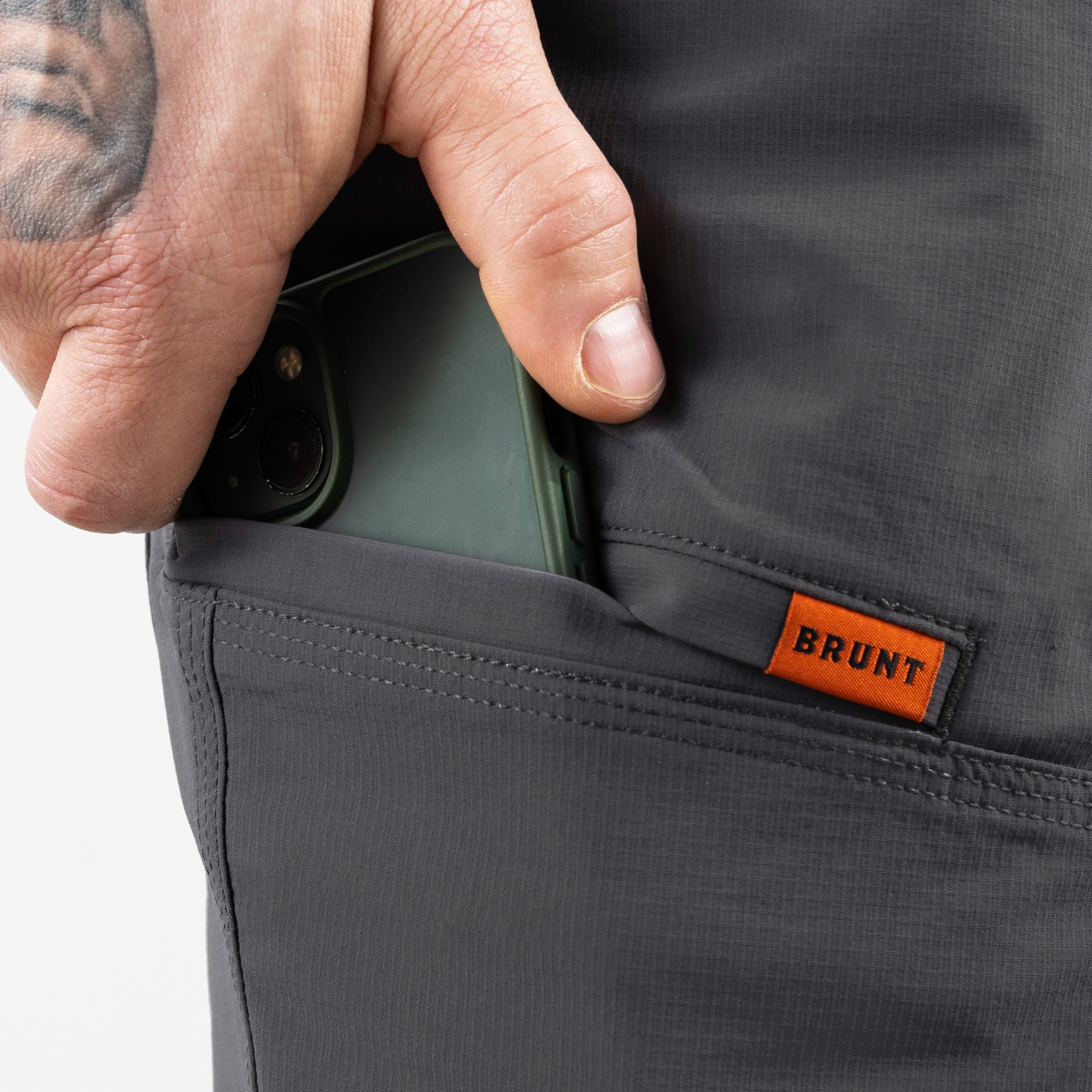 Person inserting a phone into a pocket of dark gray pants with 'BRUNT' branding.