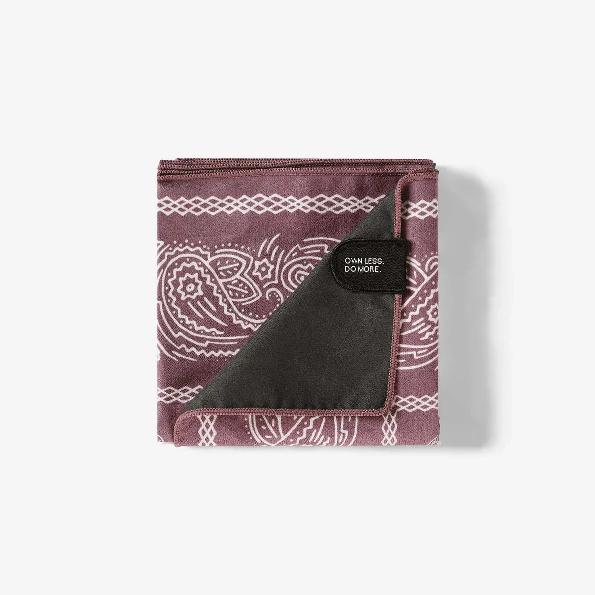 A folded yoga blanket with a dusty rose and white paisley pattern, featuring a dark charcoal gray textured underside and a black brand patch that reads 'OWNLESS. DO MORE.