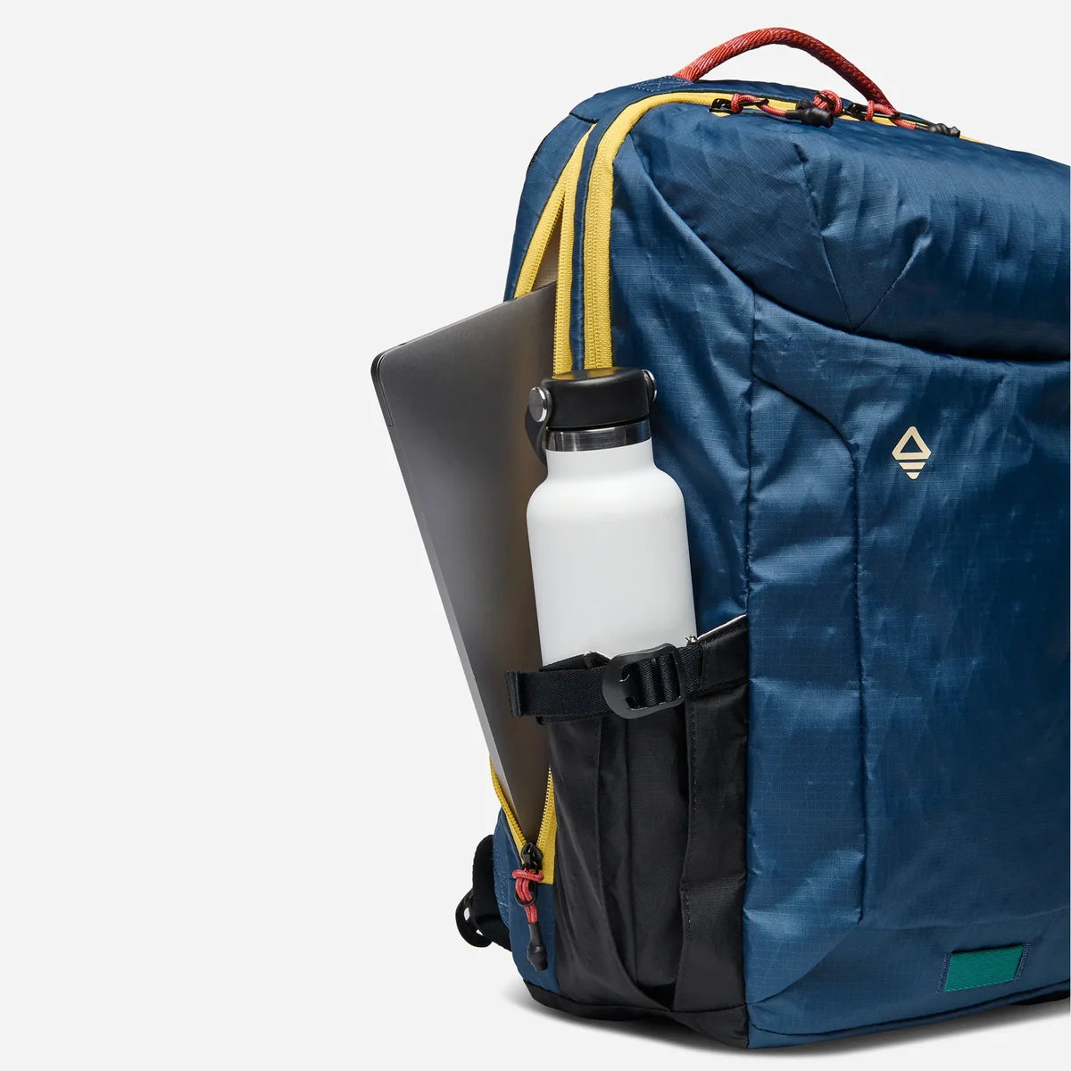 A blue technical backpack with yellow and black accents, shown with a white water bottle and a dark gray laptop in its side pockets, on a light gray background.