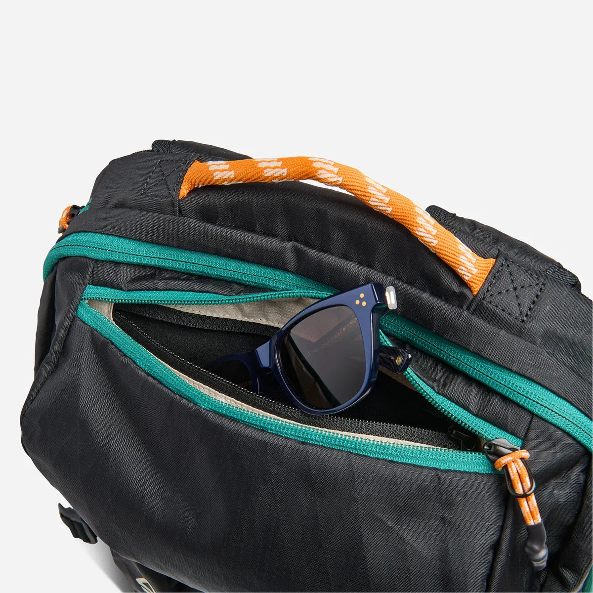 Close-up of a black backpack with teal zippers and an orange-and-white woven handle, shown with sunglasses tucked into one of the front zip pockets against a light gray background.