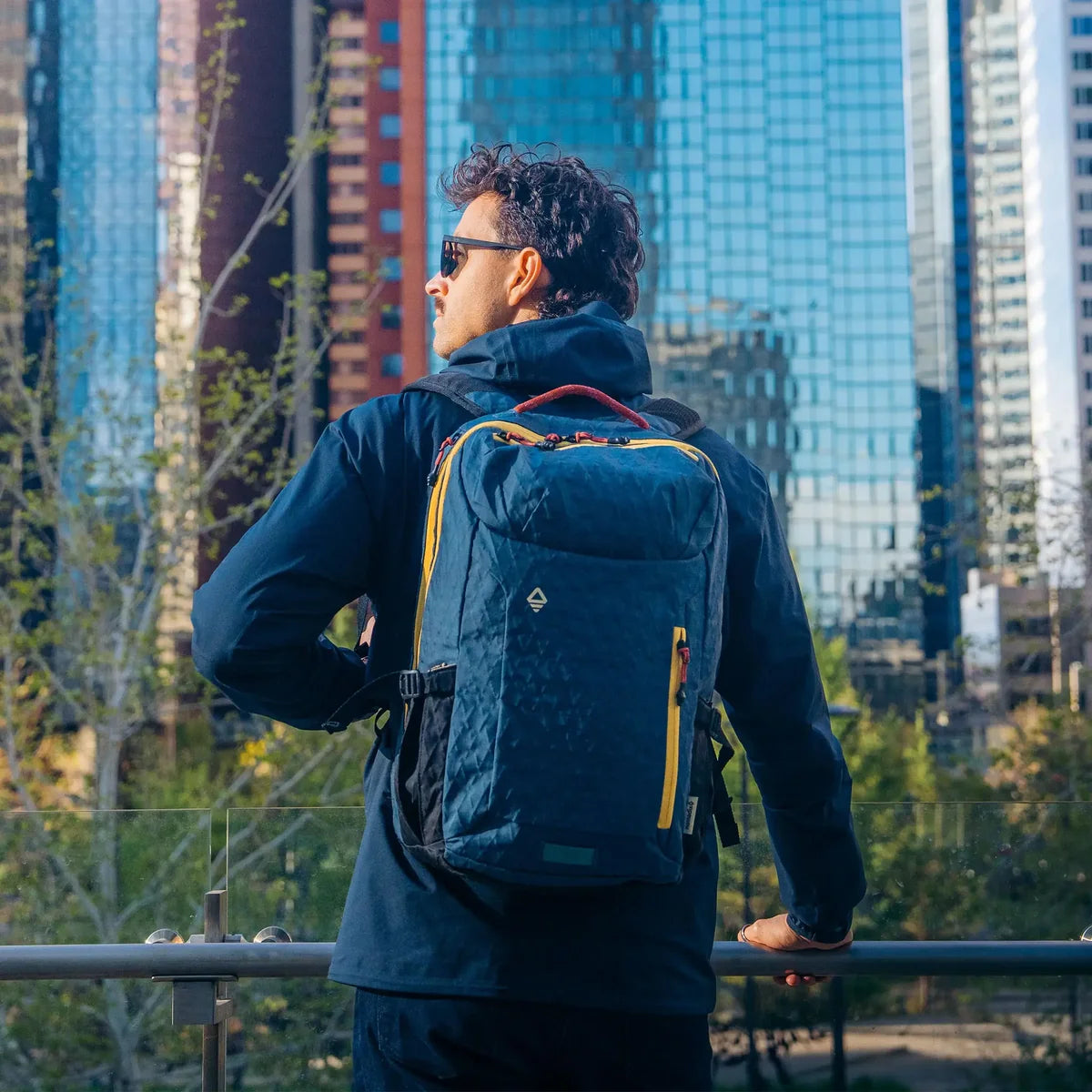 A man wearing a navy blue backpack with yellow accents, photographed from behind while looking out over a city skyline with tall glass buildings.
