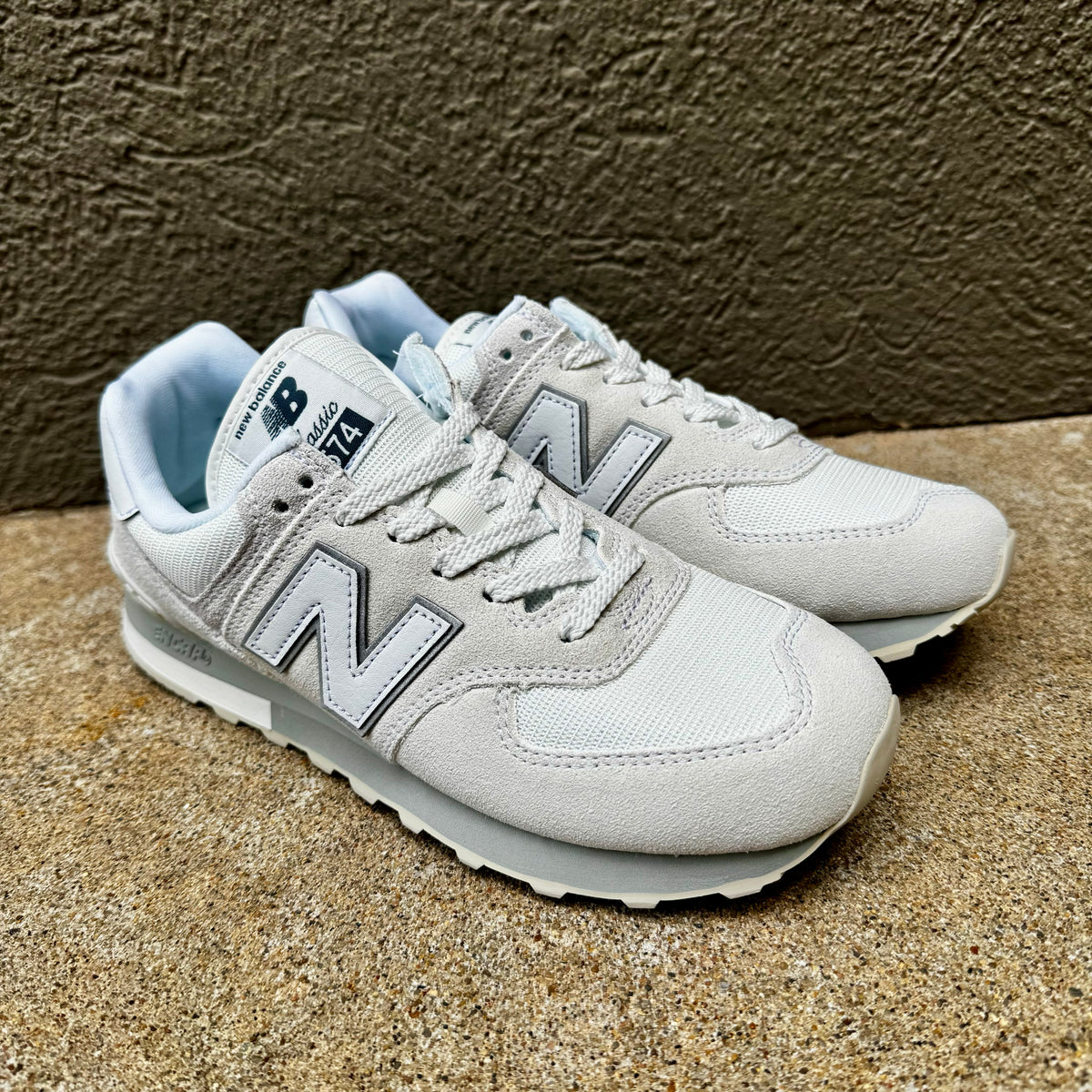 A pair of New Balance 574 sneakers in light gray and white, featuring the signature 'N' logo, lace-up closure, and light blue lining, set against a textured brown background.