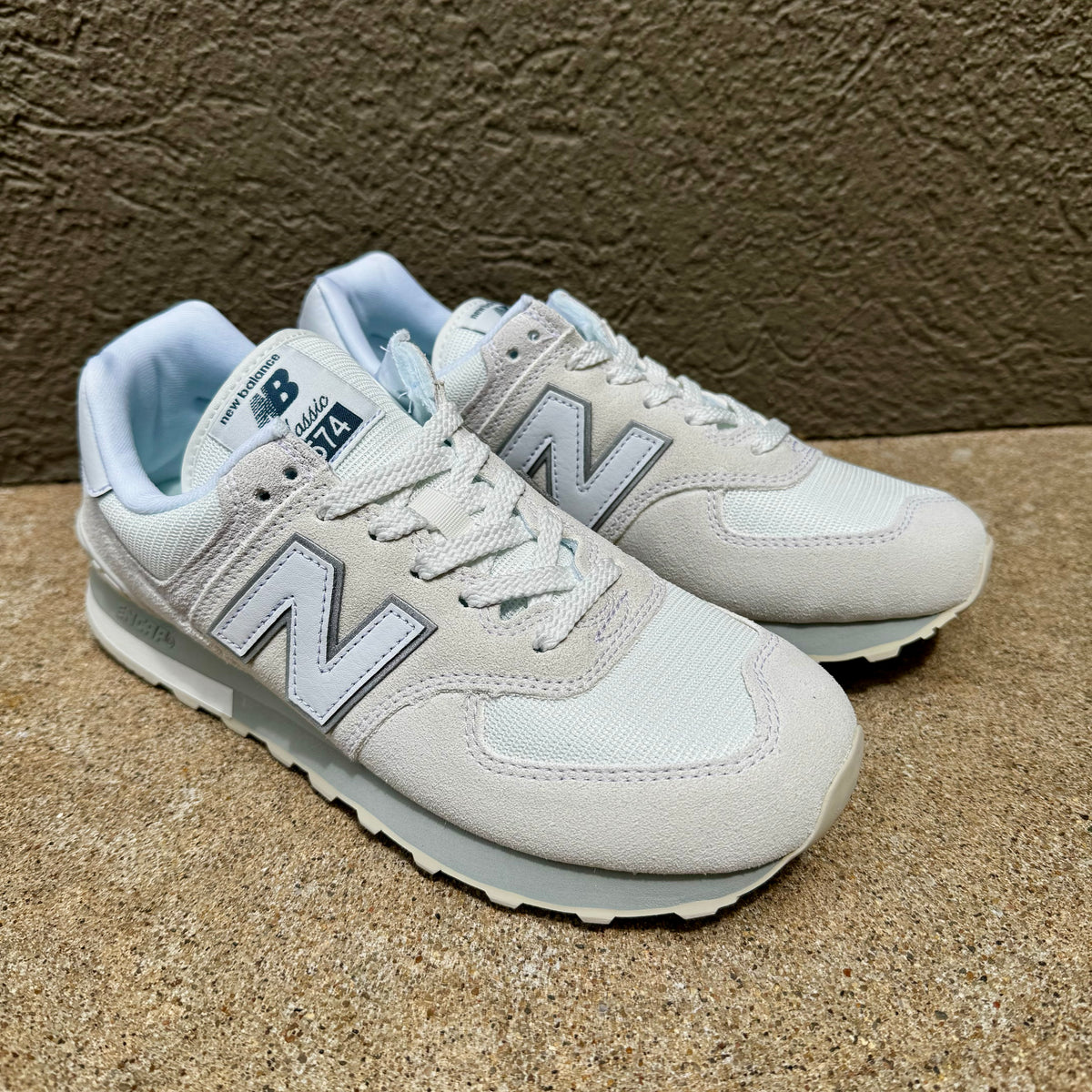 A pair of New Balance 574 sneakers in light gray and white, displayed on a textured concrete surface against a brown wall.