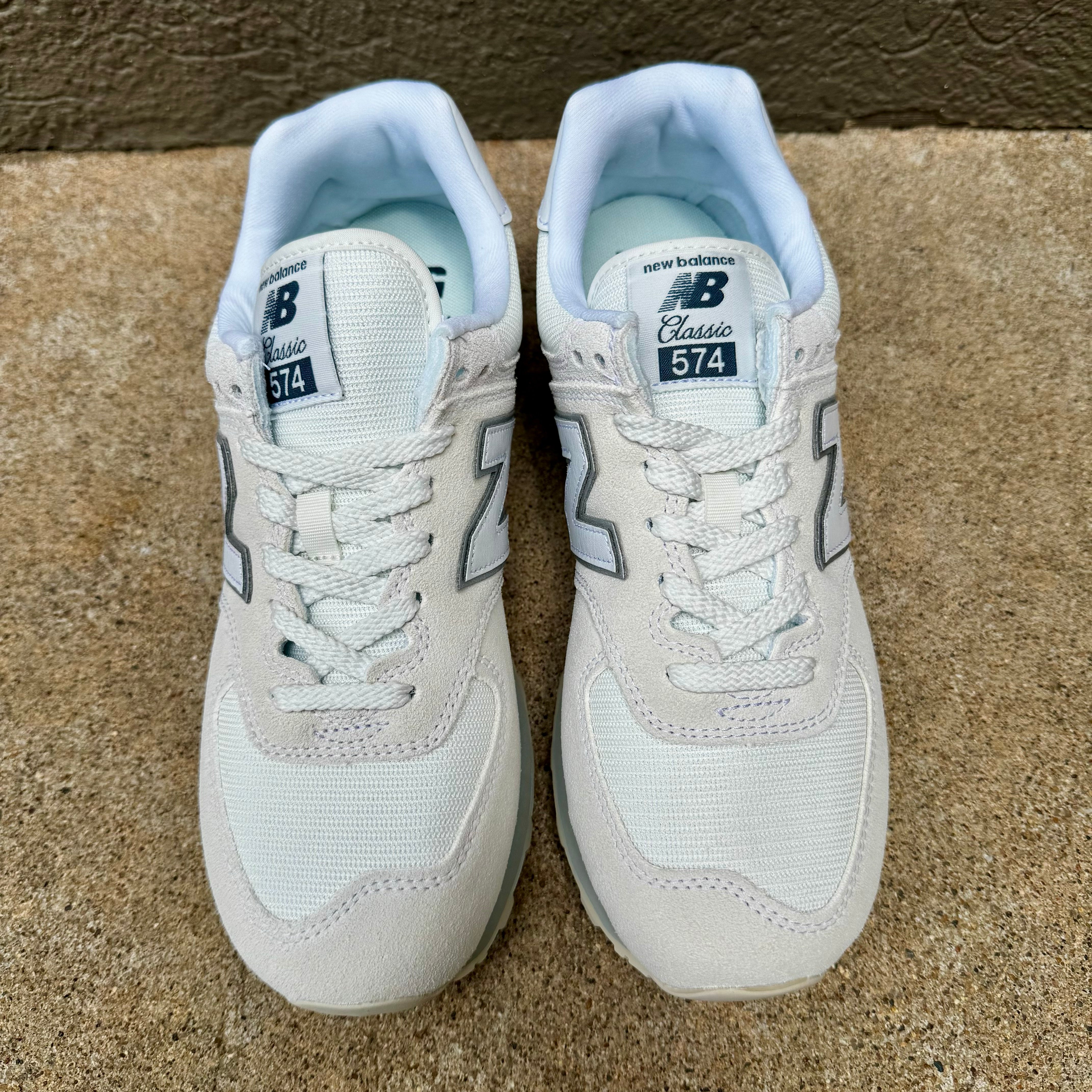 A pair of New Balance 574 Classic sneakers in white and light blue, shown from the front on a textured concrete floor.