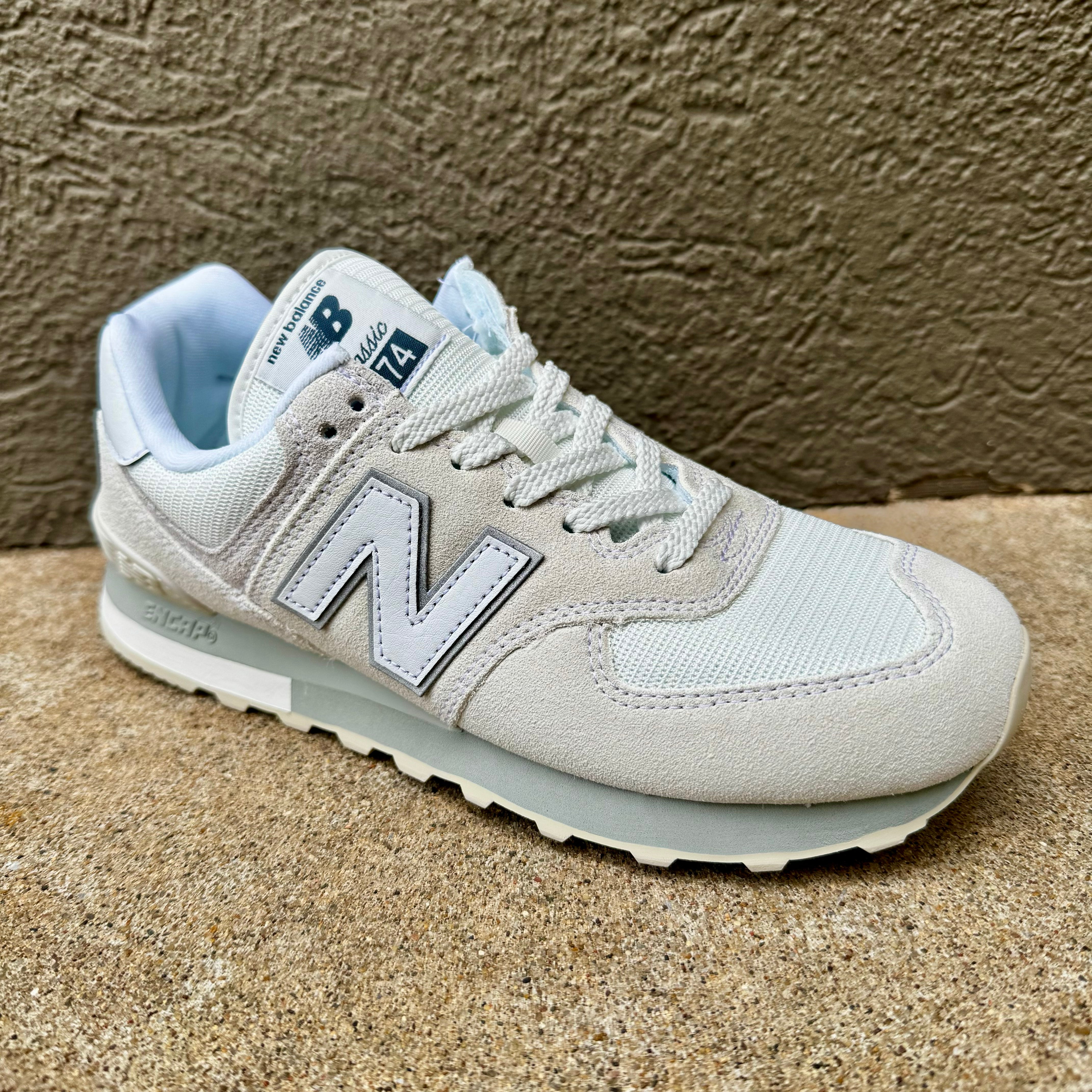 A light gray and white New Balance 574 sneaker with a light blue lining, photographed on a textured concrete surface.