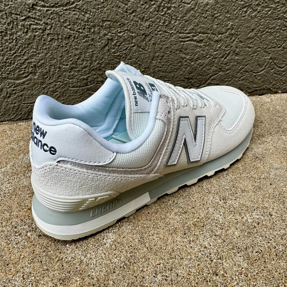 A single New Balance sneaker in white and light gray with a pale blue lining, photographed on a textured concrete surface against a stucco wall.