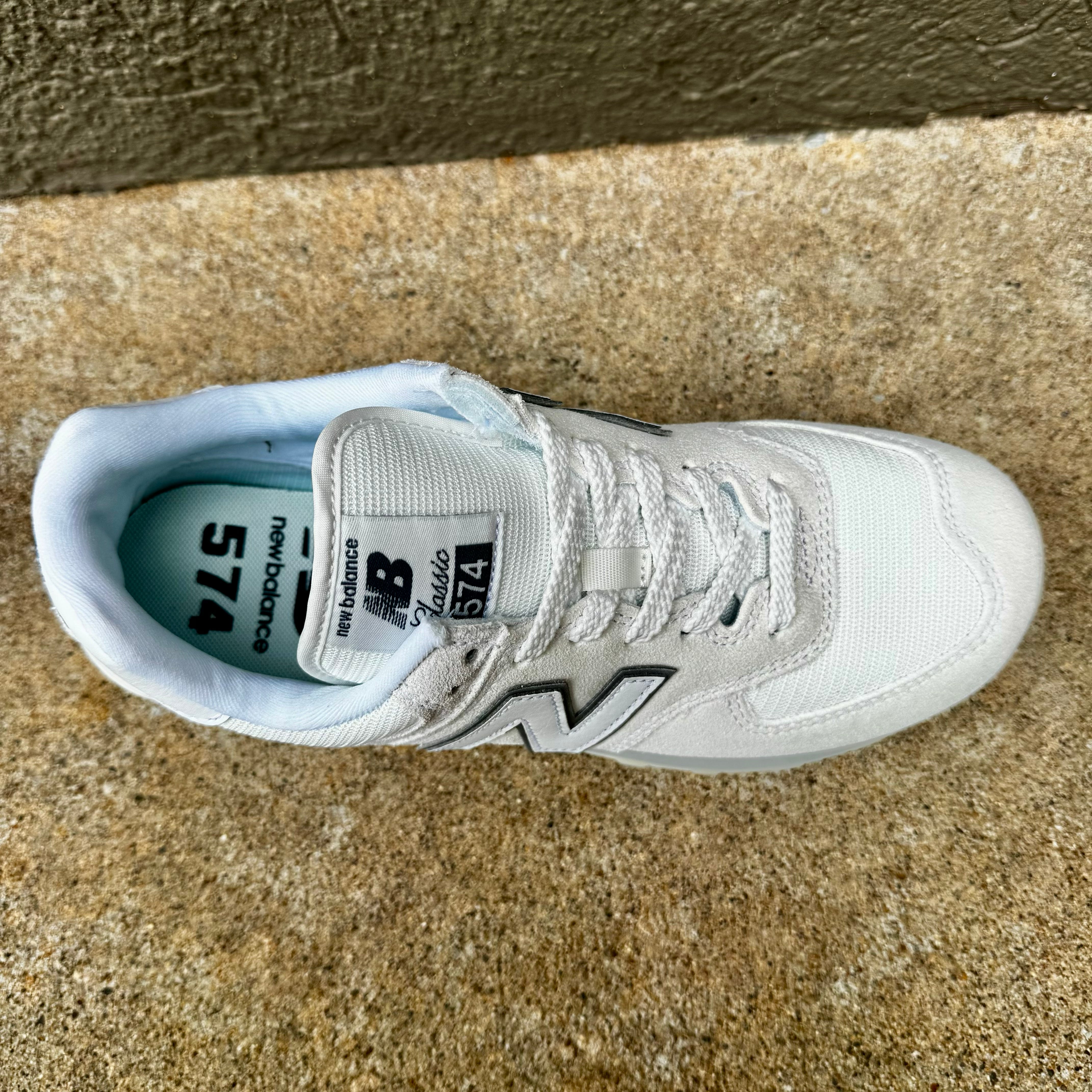 A single New Balance 574 sneaker in white and light gray, photographed from a top-down angle on a textured concrete surface.