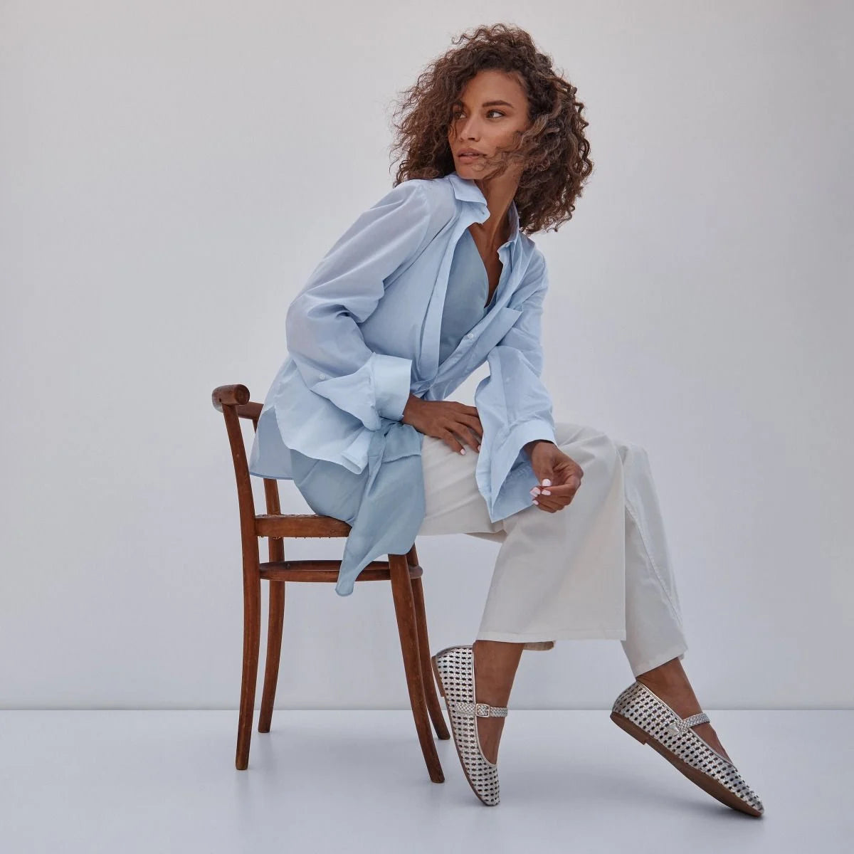 Woman sitting on a chair wearing a light blue shirt and white pants with a white background