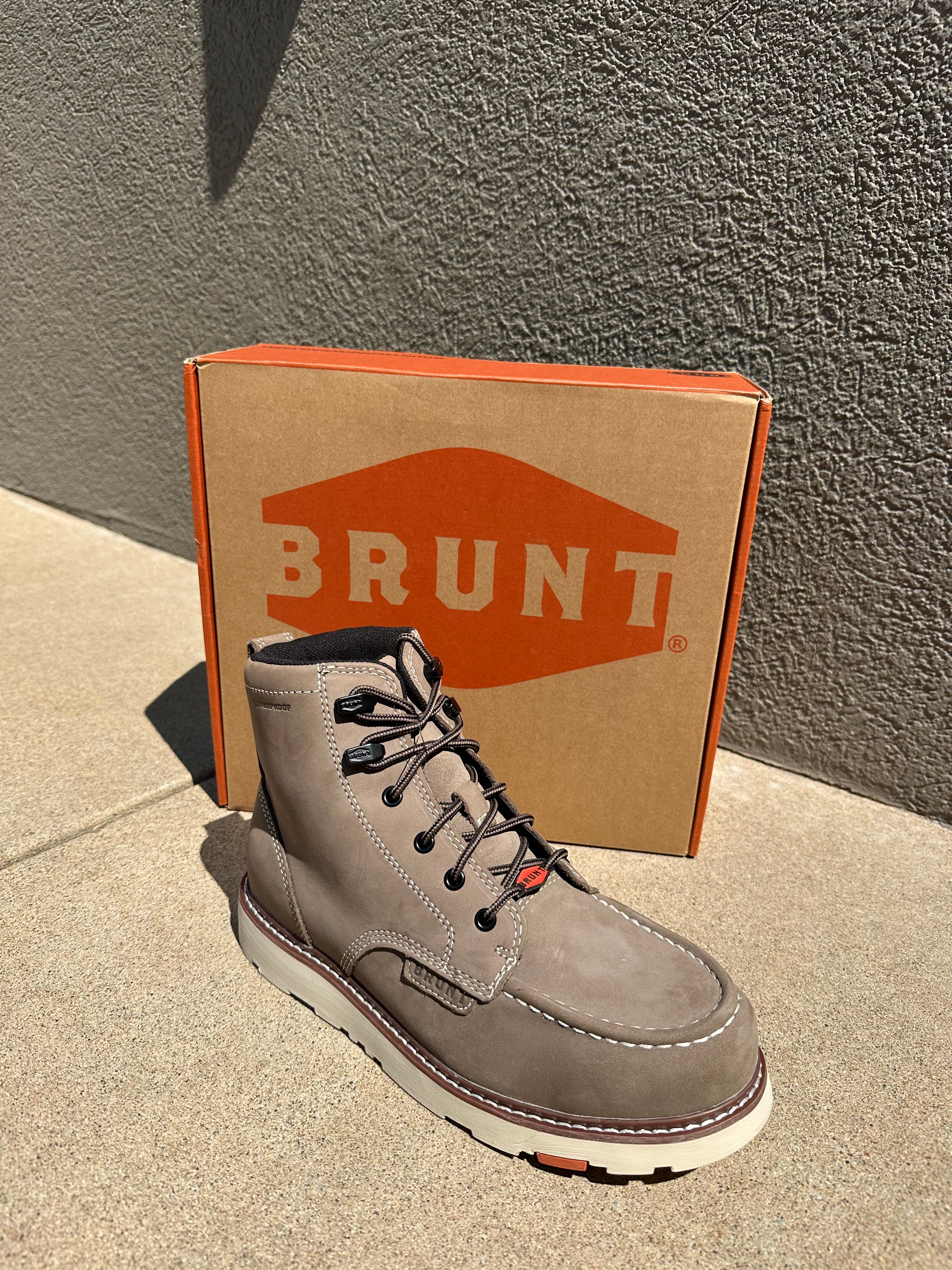 A gray suede lace-up work boot positioned in front of its cardboard packaging box, which features the orange and white BRUNT logo. The scene is set outdoors on a concrete surface against a textured gray wall under bright sunlight.