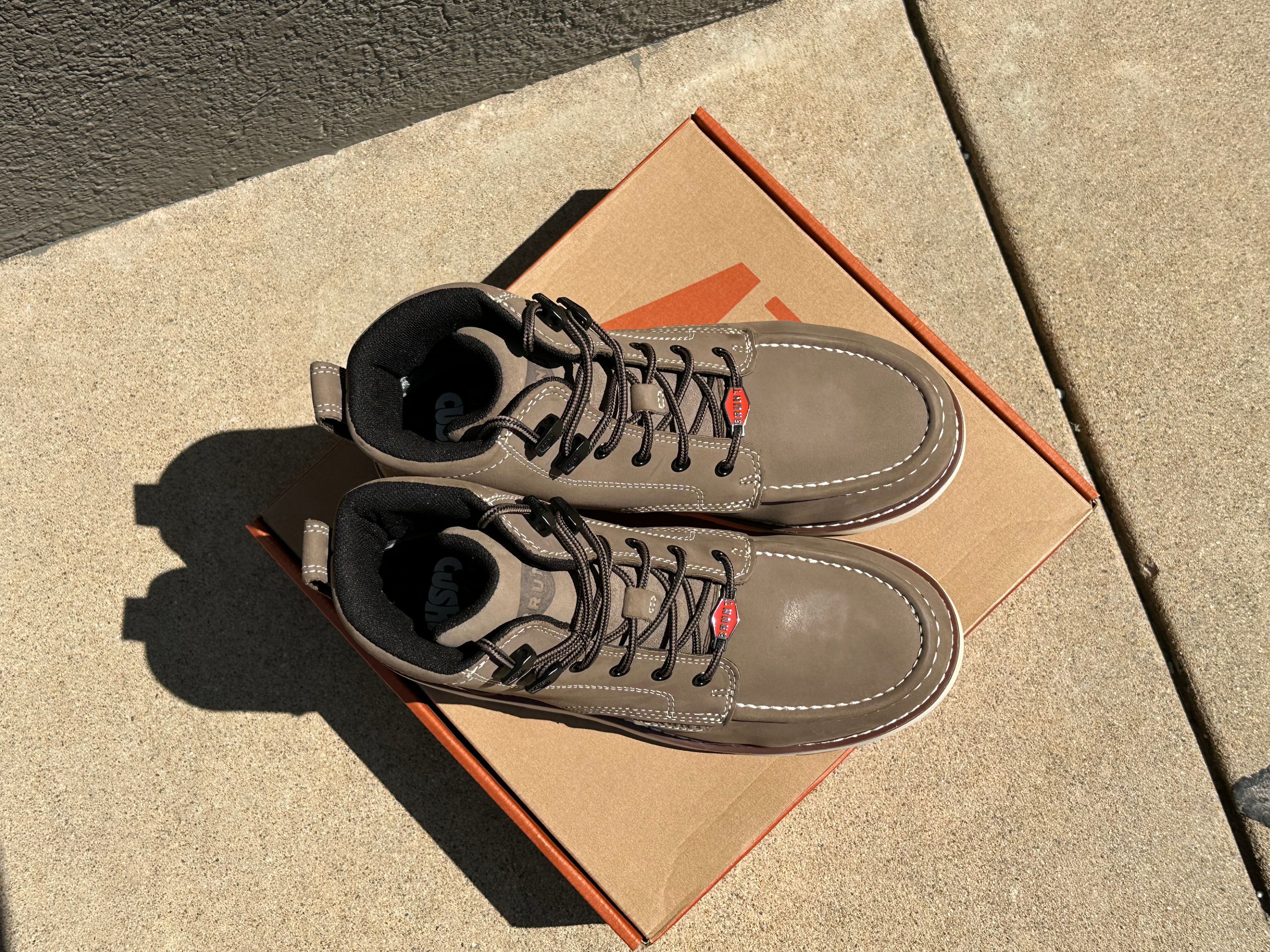 A pair of taupe-brown leather lace-up boots resting on top of their brown cardboard shoebox, placed on a concrete sidewalk under bright sunlight.