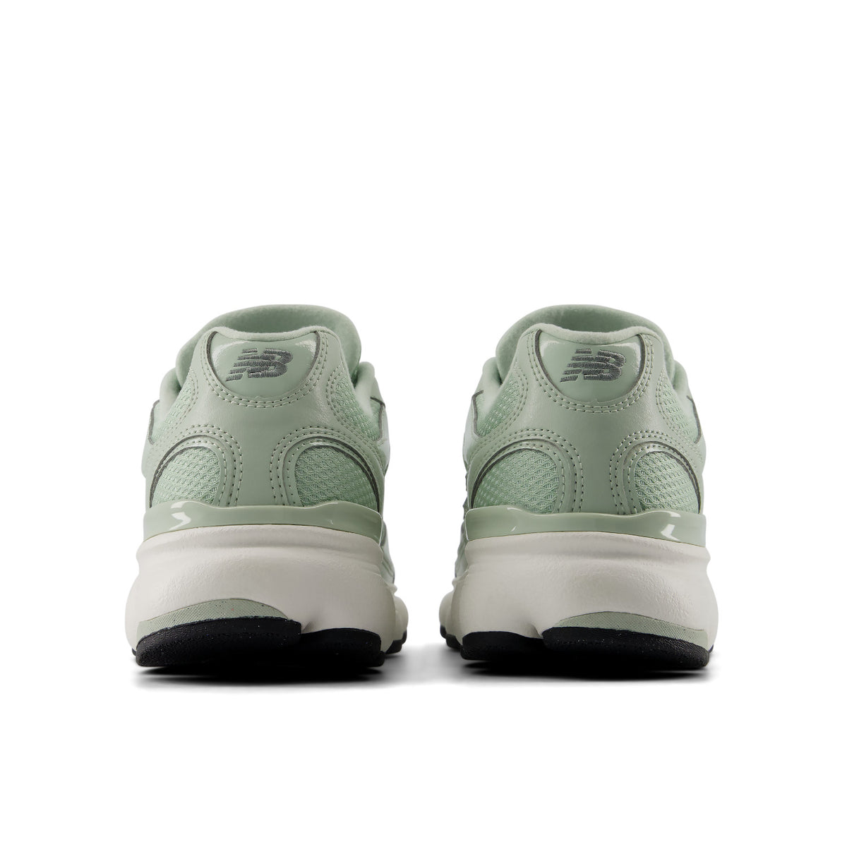 A pair of New Balance sneakers shown from the back, featuring a light sage green upper, a thick cream midsole, and a black outsole, set against a plain white background.