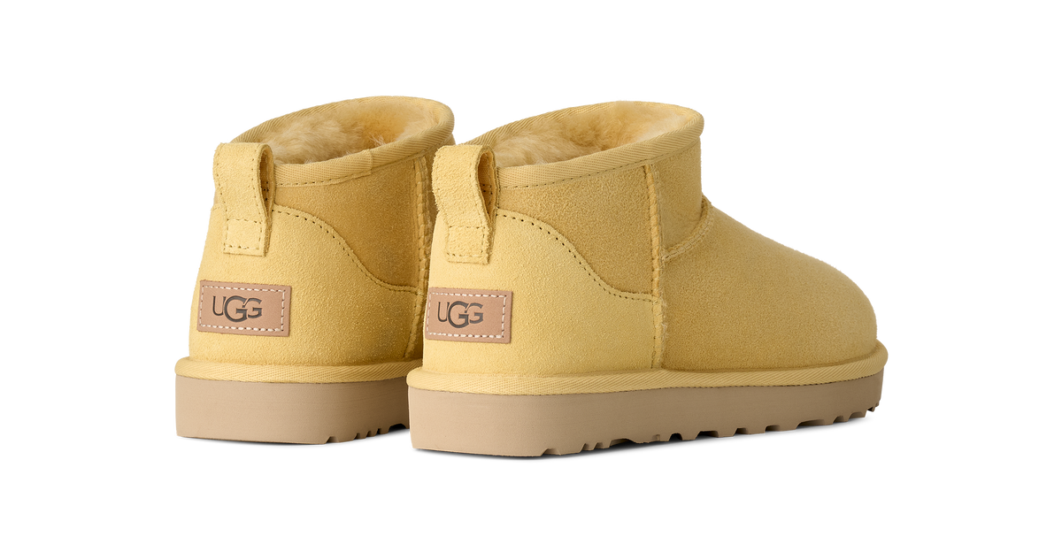 A pair of UGG Mini Slipper boots in a warm mustard yellow suede with a tan sole and a beige pull tab featuring the UGG logo.