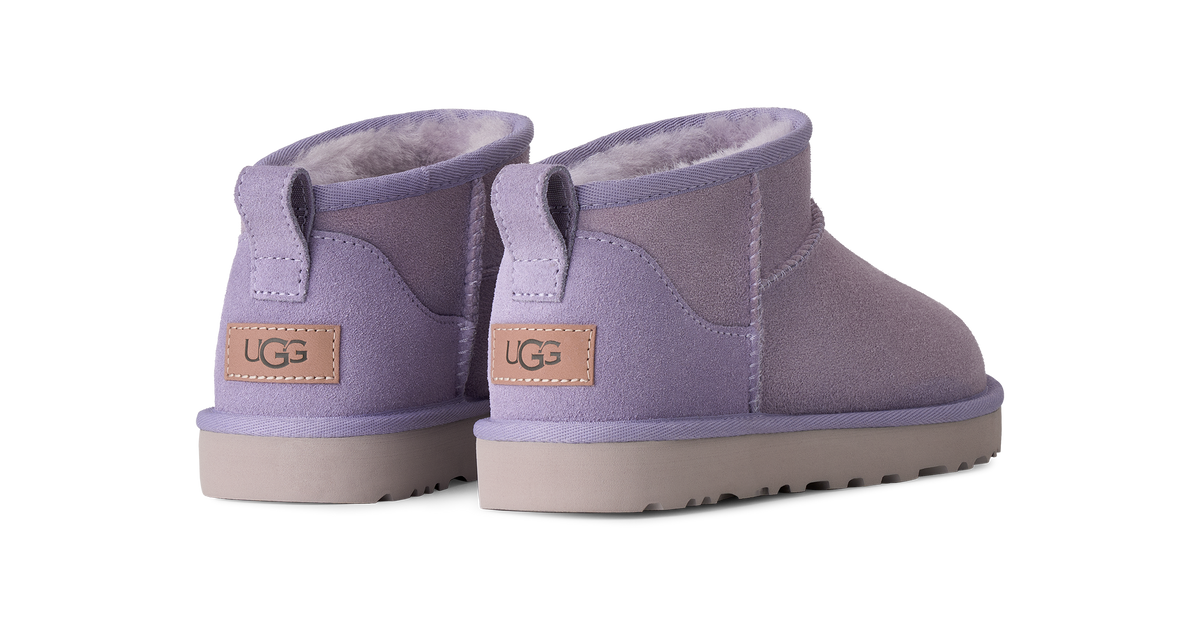 A pair of UGG mini ankle boots in a dusty lavender purple suede, featuring a tan leather UGG logo patch on the back heel and a light gray sole, shown against a solid white background.