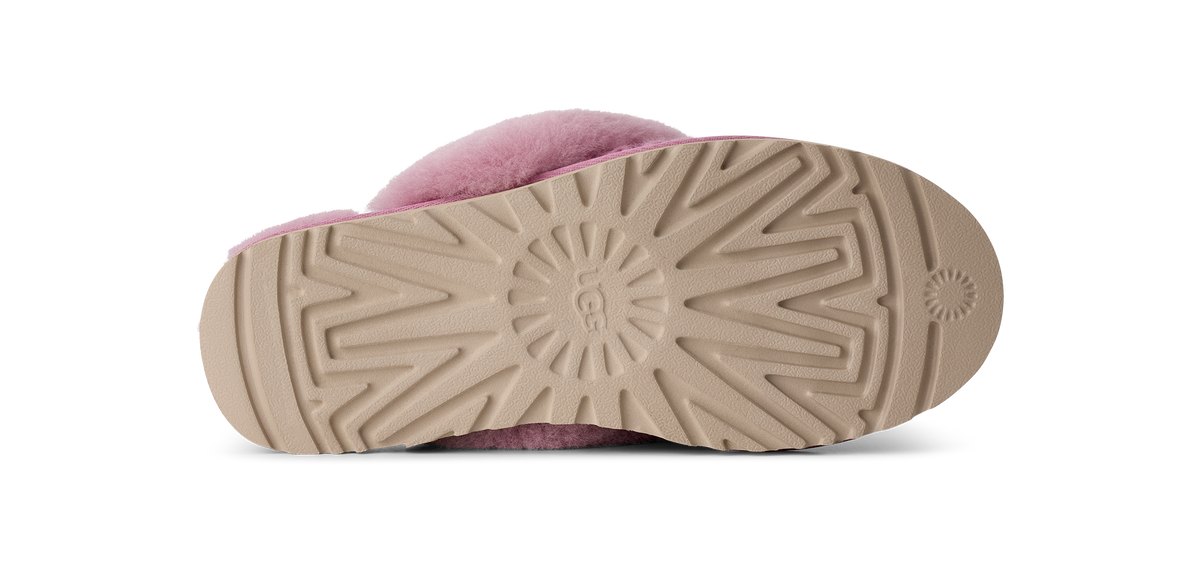 The bottom sole of a pink fuzzy slipper, showing a beige treaded rubber outsole with a circular UGG logo embossed in the center, photographed on a white background.