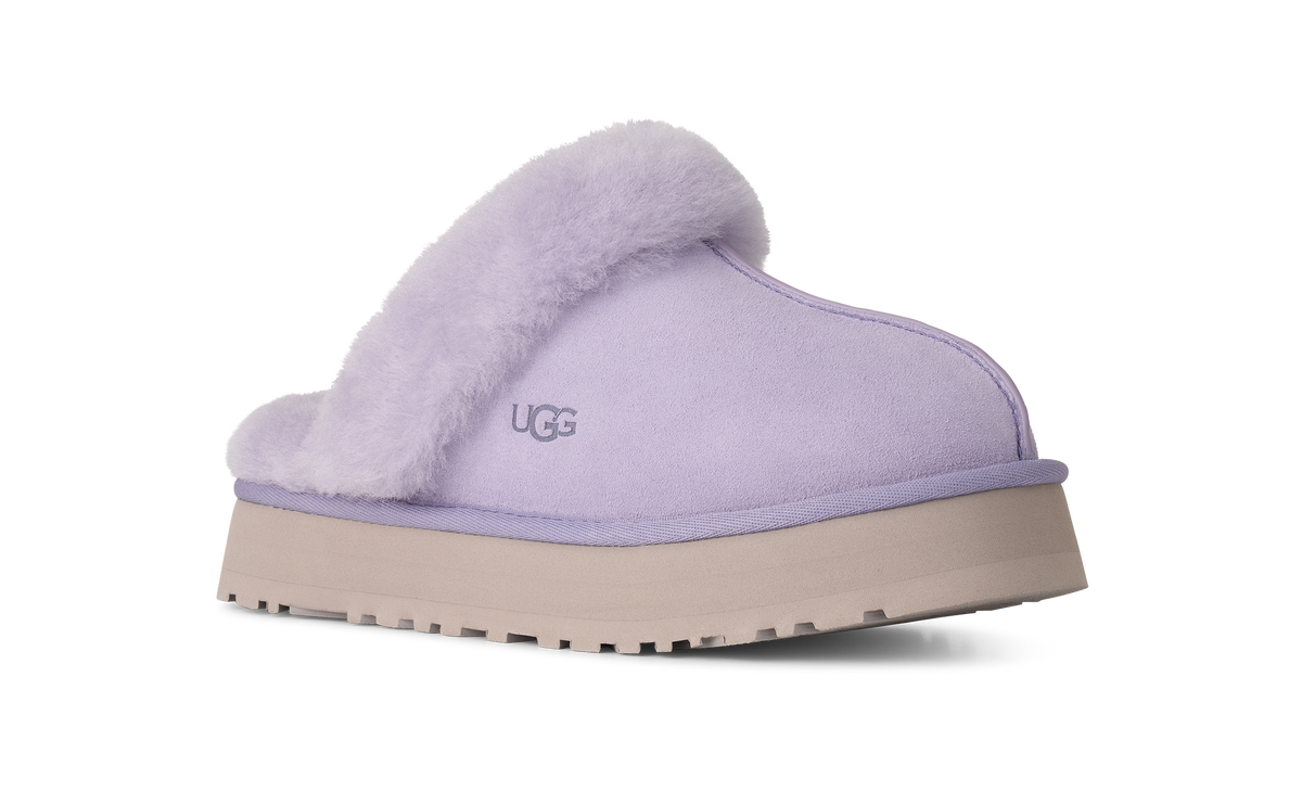 A single UGG slip-on mule slipper in light lavender suede with a fluffy lavender faux-fur collar and a beige platform sole, shown from a side profile on a white background.