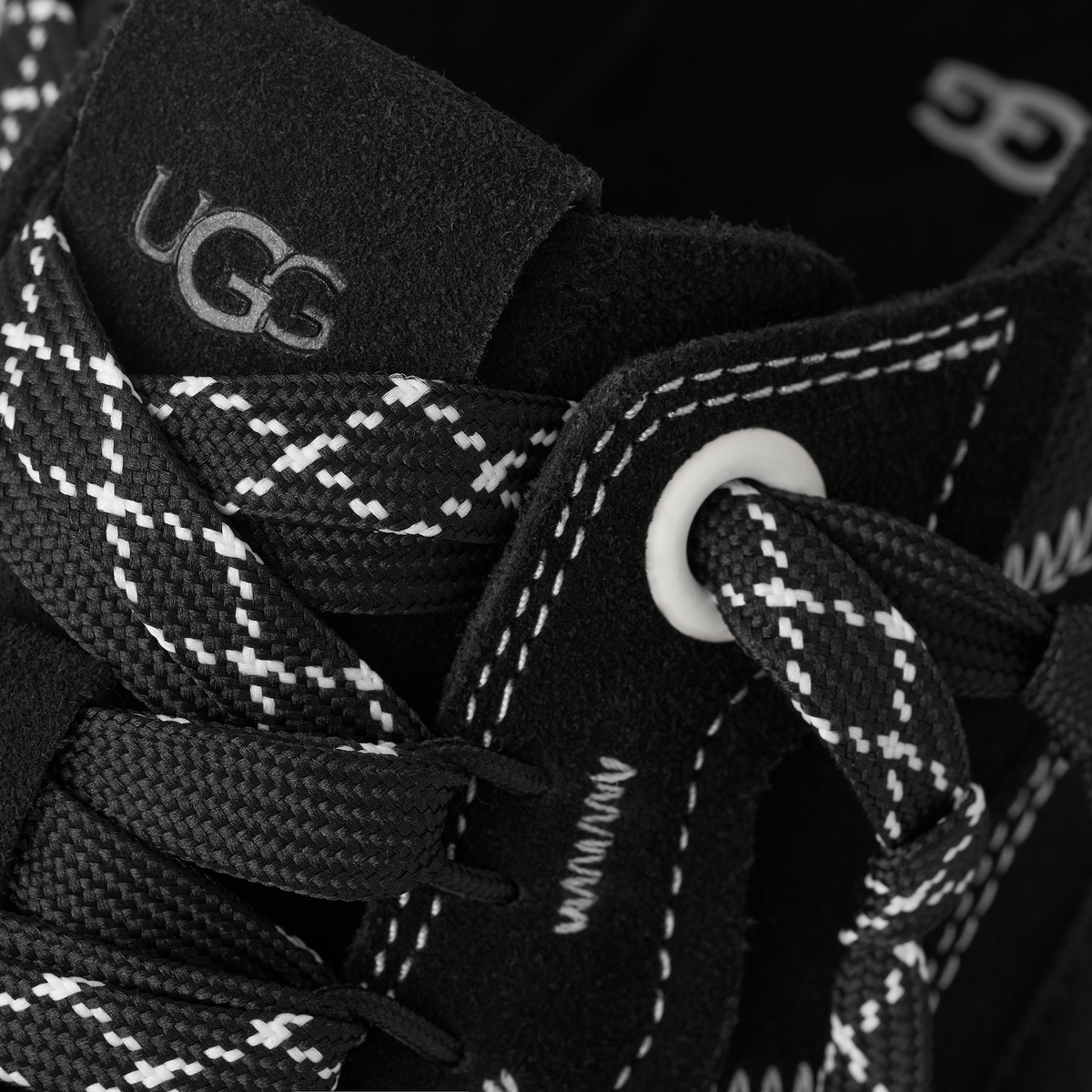 Close-up of a black UGG sneaker with white and black patterned laces, white contrast stitching, and a white UGG logo on the tongue label.