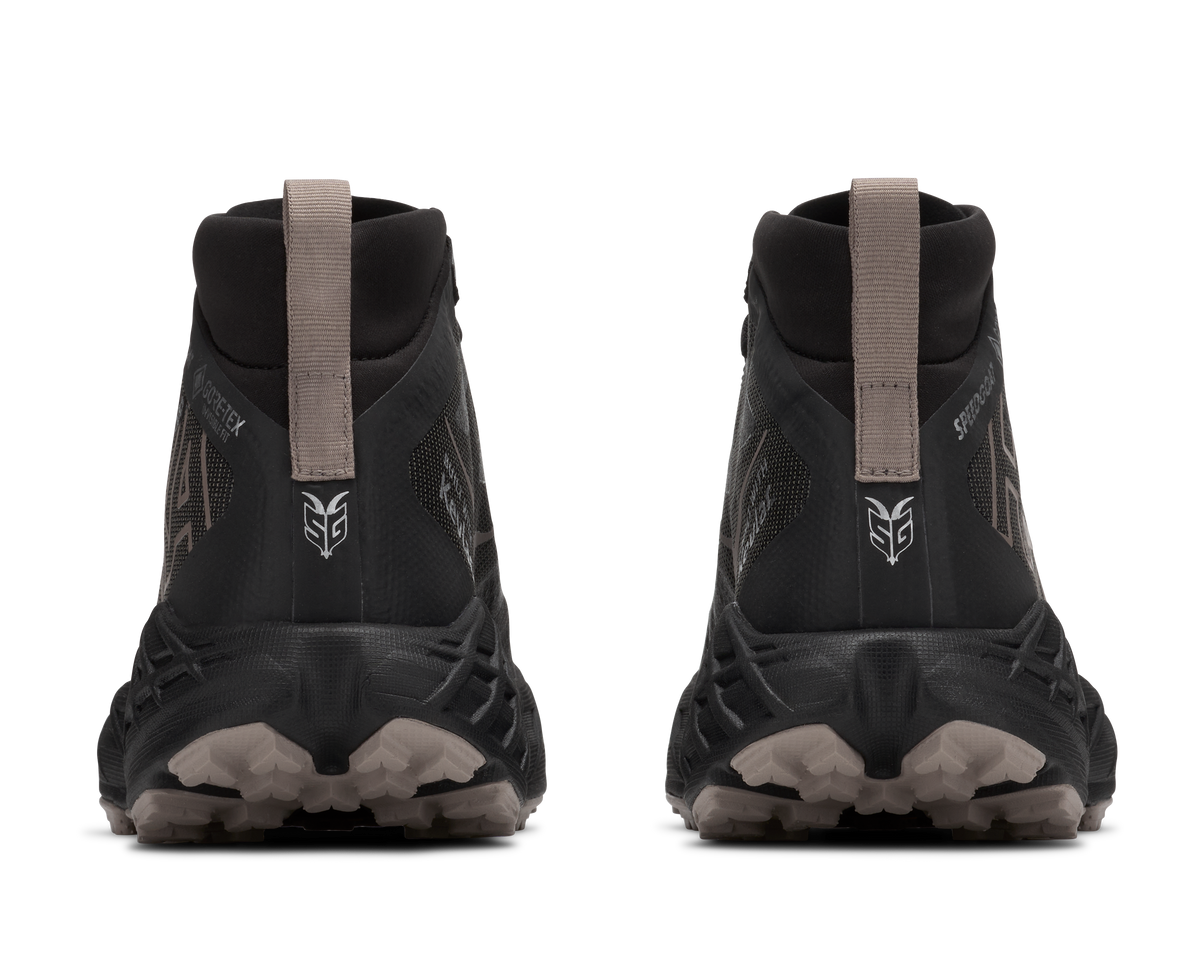 A pair of black mid-cut trail running shoes shown from the rear, featuring a pull tab, textured black and gray soles, and a white stylized logo on the heel.