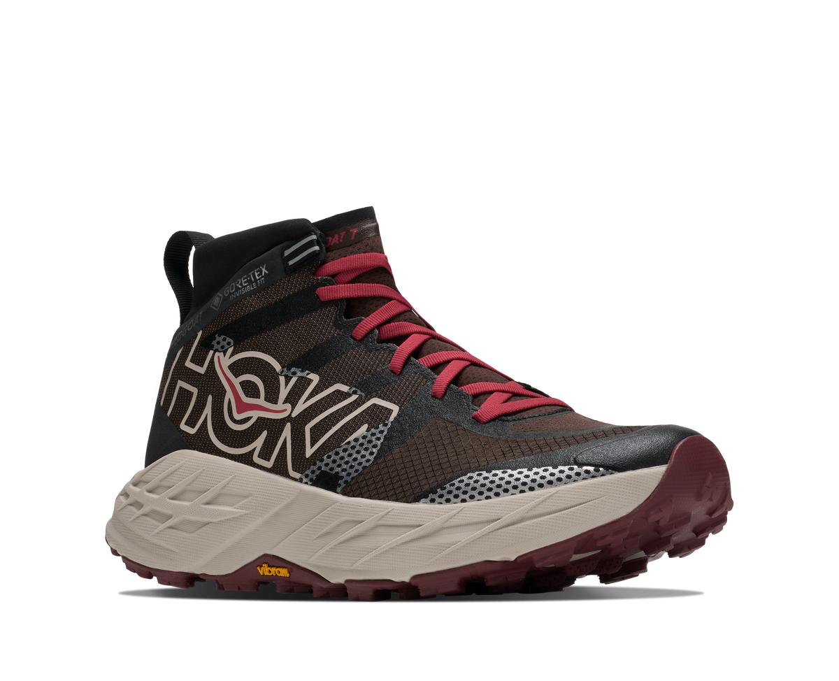 A Hoka trail running shoe in dark brown and black mesh with red laces and a beige midsole, shown from a side profile on a white background.