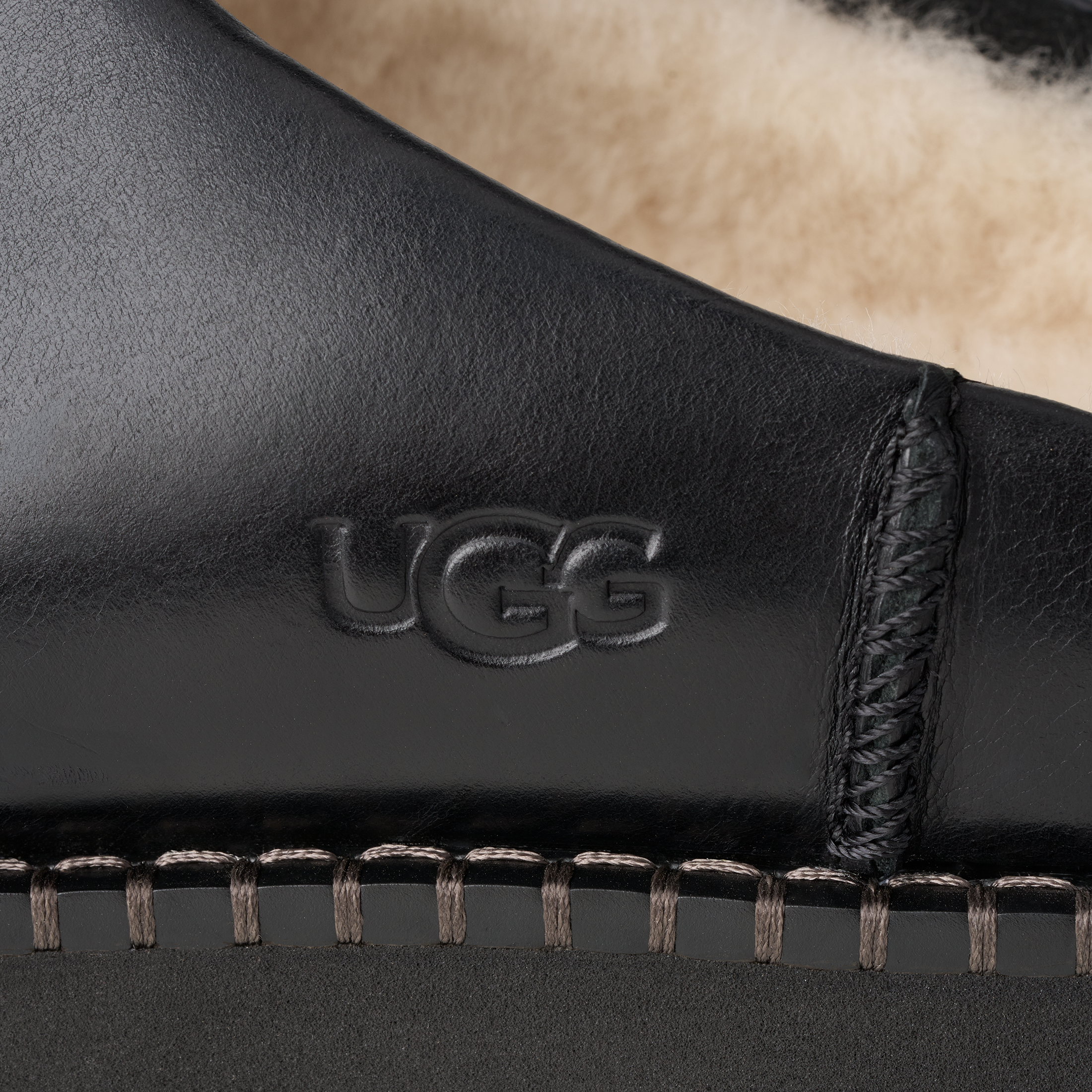 Close-up detail of a black leather UGG boot showing the embossed UGG logo, contrast stitching, and a beige shearling lining.