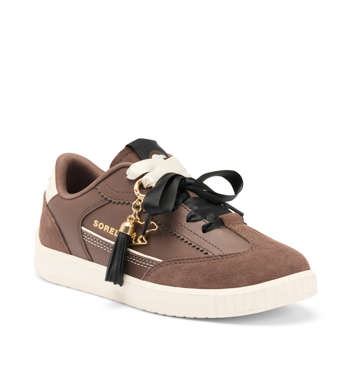 A brown suede and leather low-top sneaker with black laces, a black ribbon bow, a gold-tone tassel charm, and a cream-colored sole, shown on a white background.
