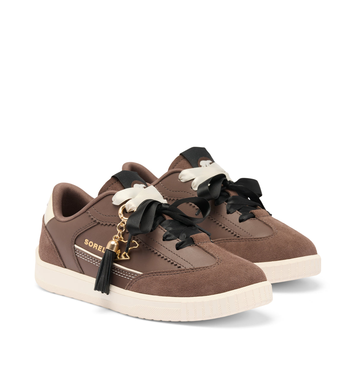 A pair of Sorel brand low-top sneakers in brown suede and leather, featuring black laces with white decorative bows, a black tassel with a gold key charm, and a cream-colored sole.