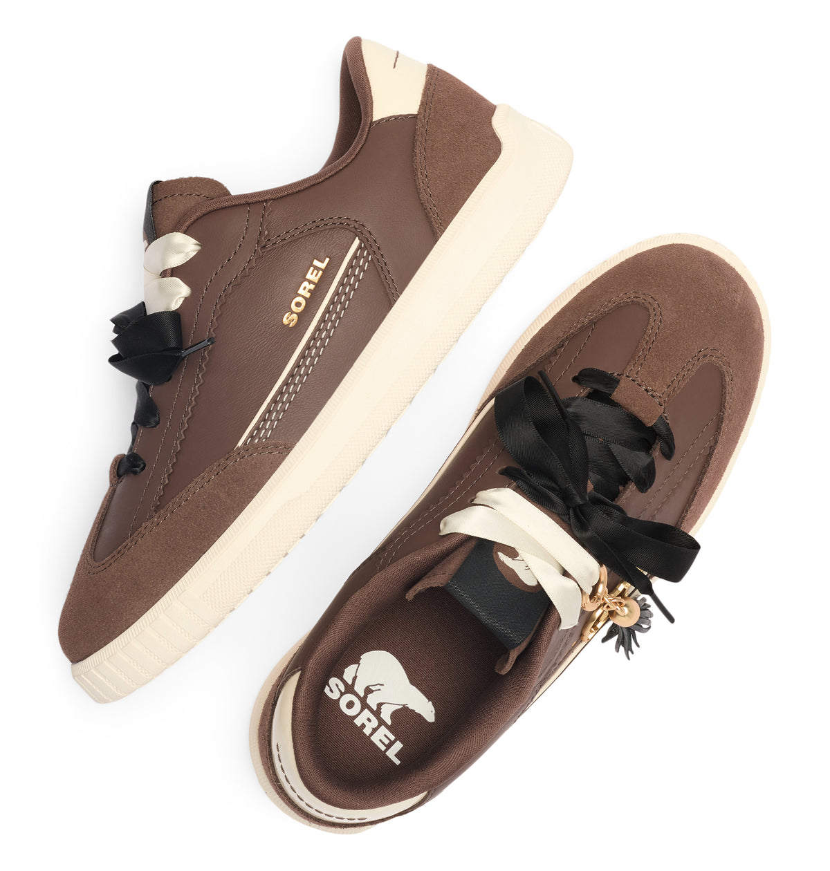 A pair of Sorel low-top sneakers in brown suede and leather, featuring cream-colored midsoles, black decorative laces with white ribbon accents, and a small gold bear charm on the laces. One shoe is shown from a side angle and the other from a top-down angle, both on a clean white background.