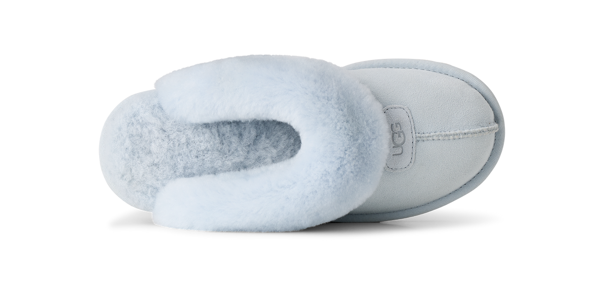 A single UGG slipper shown from the side profile against a clean white background. The slipper features a light blue suede exterior and a fluffy pale blue shearling lining, with the 'UGG' brand logo visible on a small side tab.