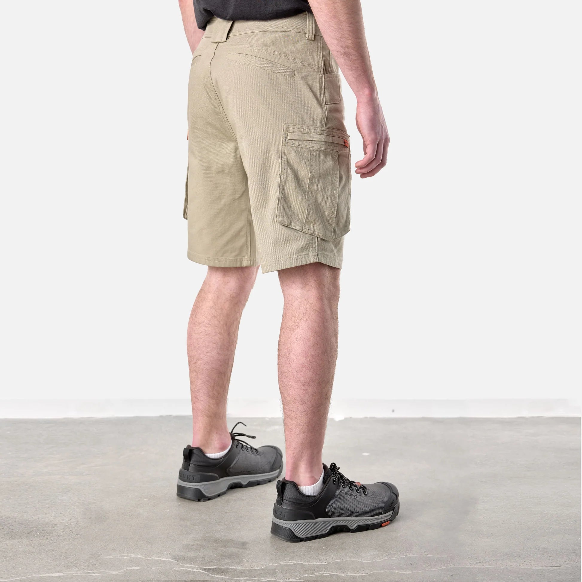 A person shown from the back wearing khaki cargo shorts, a dark shirt, and dark gray hiking shoes against a plain studio background.