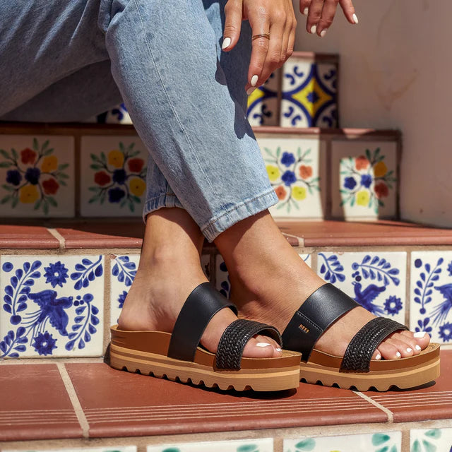 Person wearing black sandals with brown soles on a tiled floor.