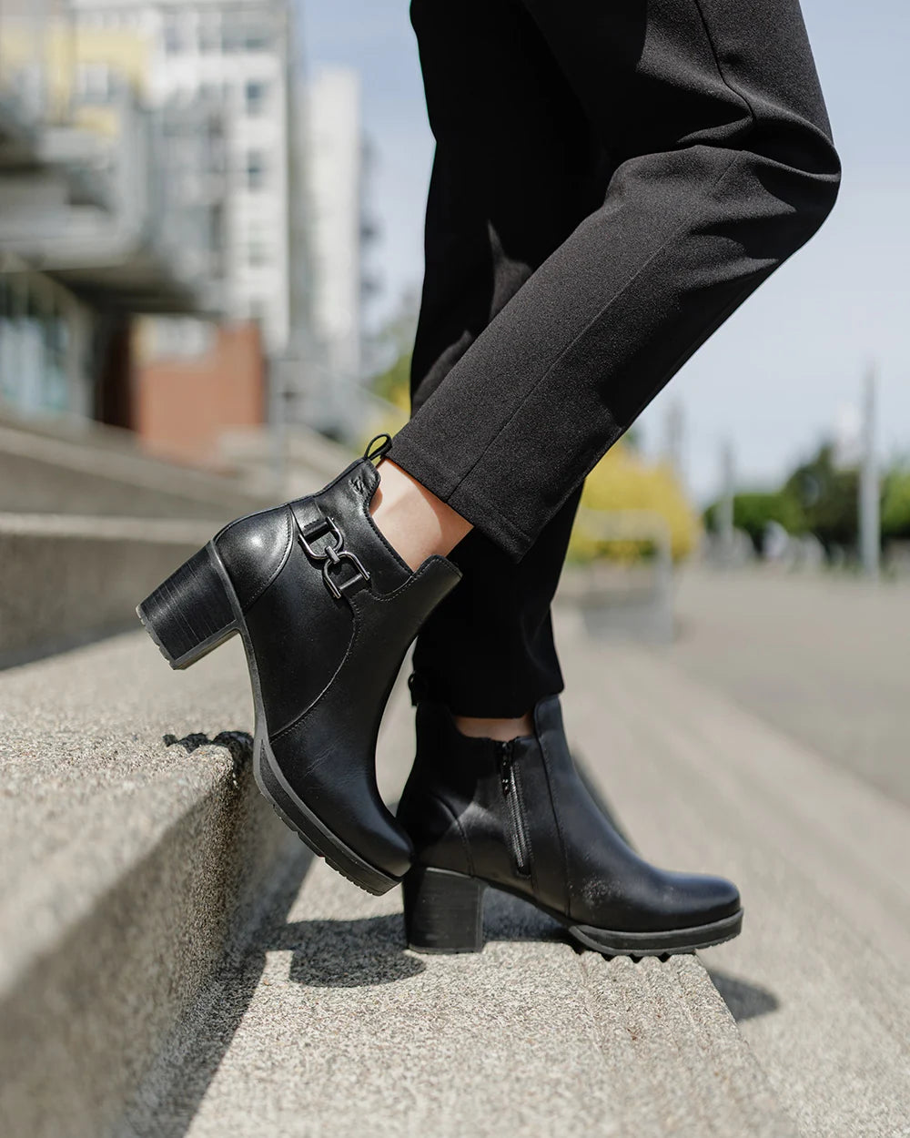 Black leather ankle boots with block heels and side buckle details, shown on a model wearing black pants while standing on concrete steps outdoors.