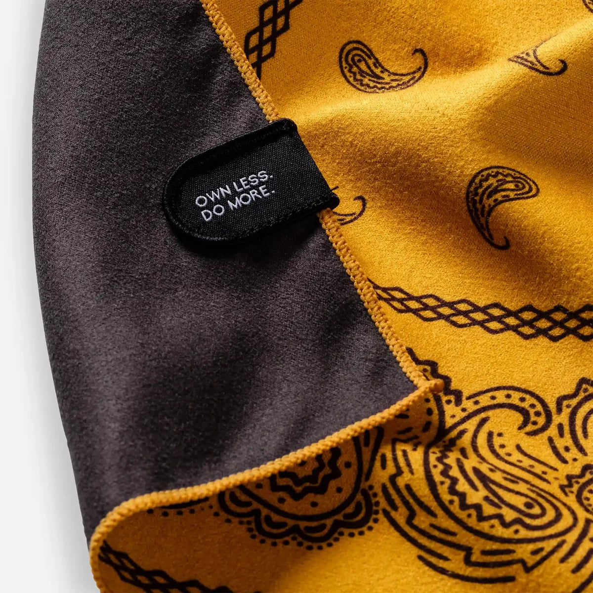 Close-up of a folded fabric item showing a mustard yellow side with black paisley and geometric line patterns, a dark charcoal gray side, and a black fabric label sewn onto the edge that reads 'OWNLESS. DO MORE.