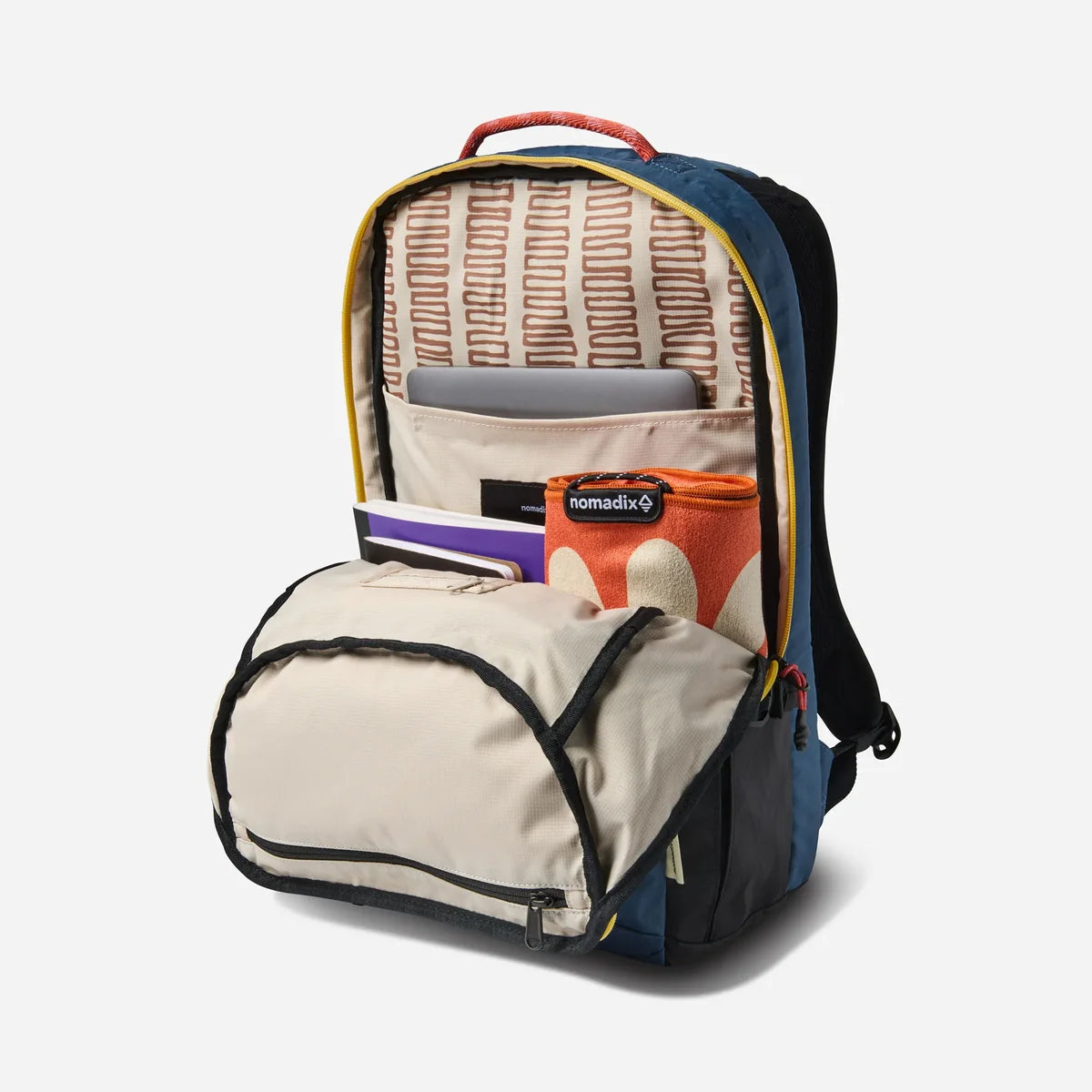 A navy blue and beige backpack shown open, revealing its interior compartments, yellow piping, and a patterned lining. A matching beige zippered pouch is placed in front, and a small orange and white patterned item is tucked inside.