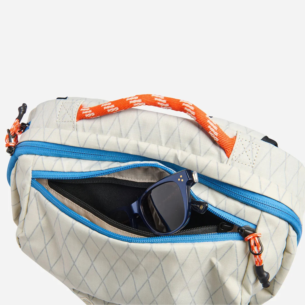A close-up of a light beige quilted bag with a blue zipper, an orange webbed handle, and an open front pocket containing blue sunglasses, on a light gray background.