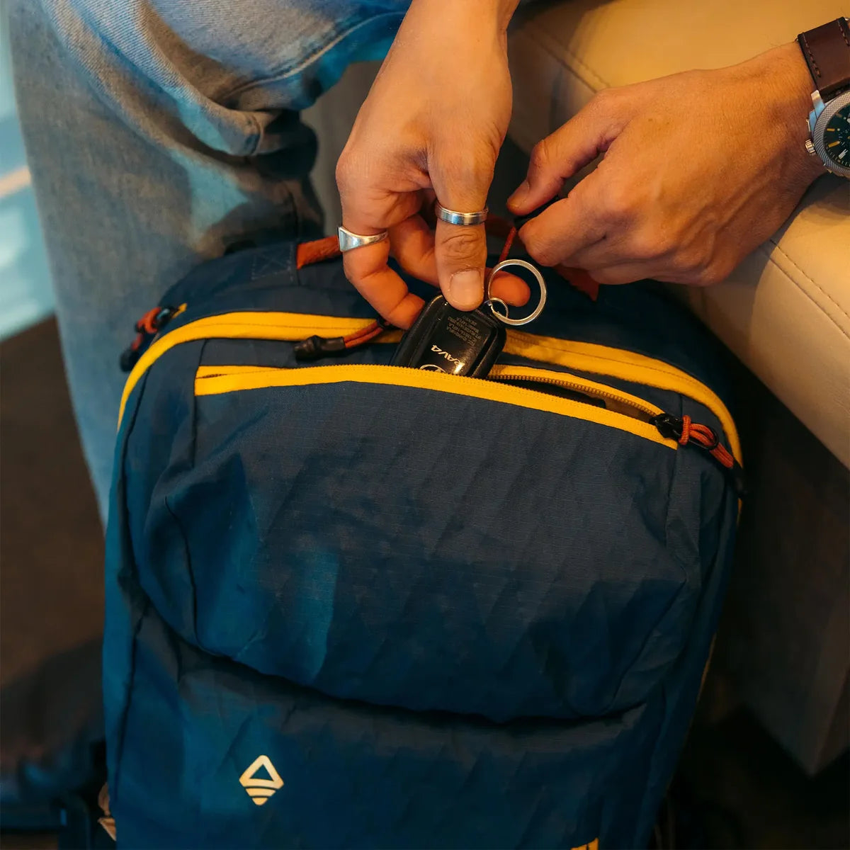 A close-up of a navy blue backpack with yellow accents, being held by a person whose hands and jeans are visible. The person is inserting a black key fob into a zippered pocket of the bag.