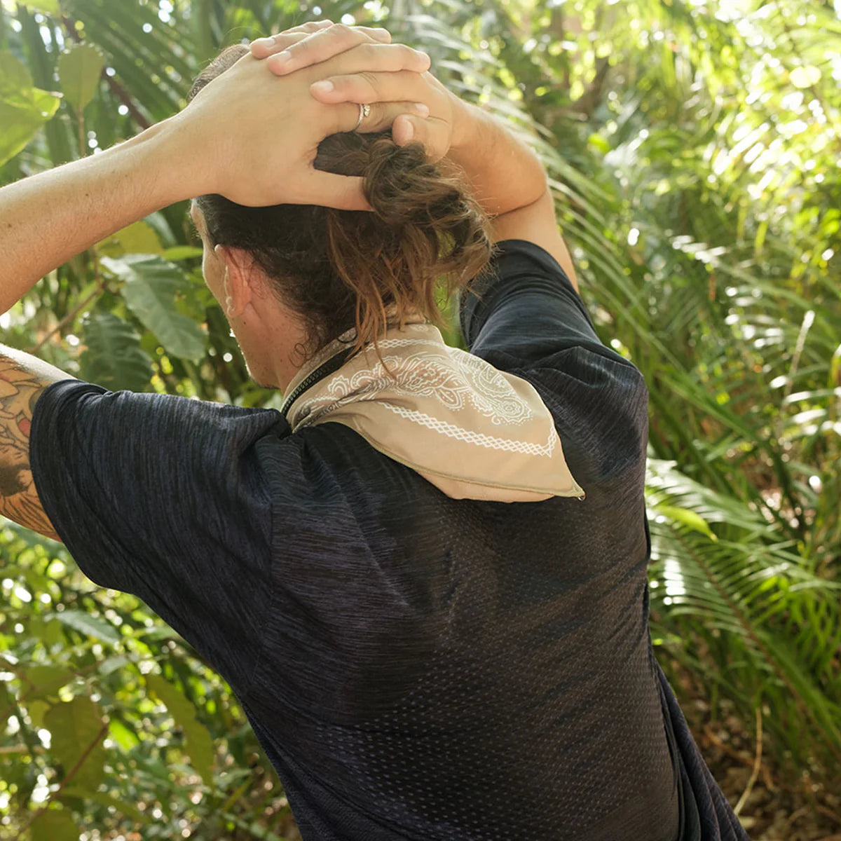A person outdoors in a lush green environment, seen from behind, wearing a dark textured athletic t-shirt and a beige patterned neck gaiter.